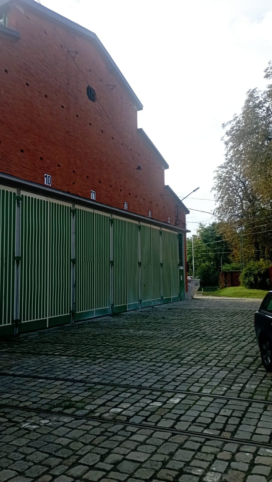 Ceglany budynek z zielonymi, numerowanymi bramami w Janikowie. Brukowana droga prowadzi wzdłuż fasady. Widoczne drzewa i fragment samochodu.