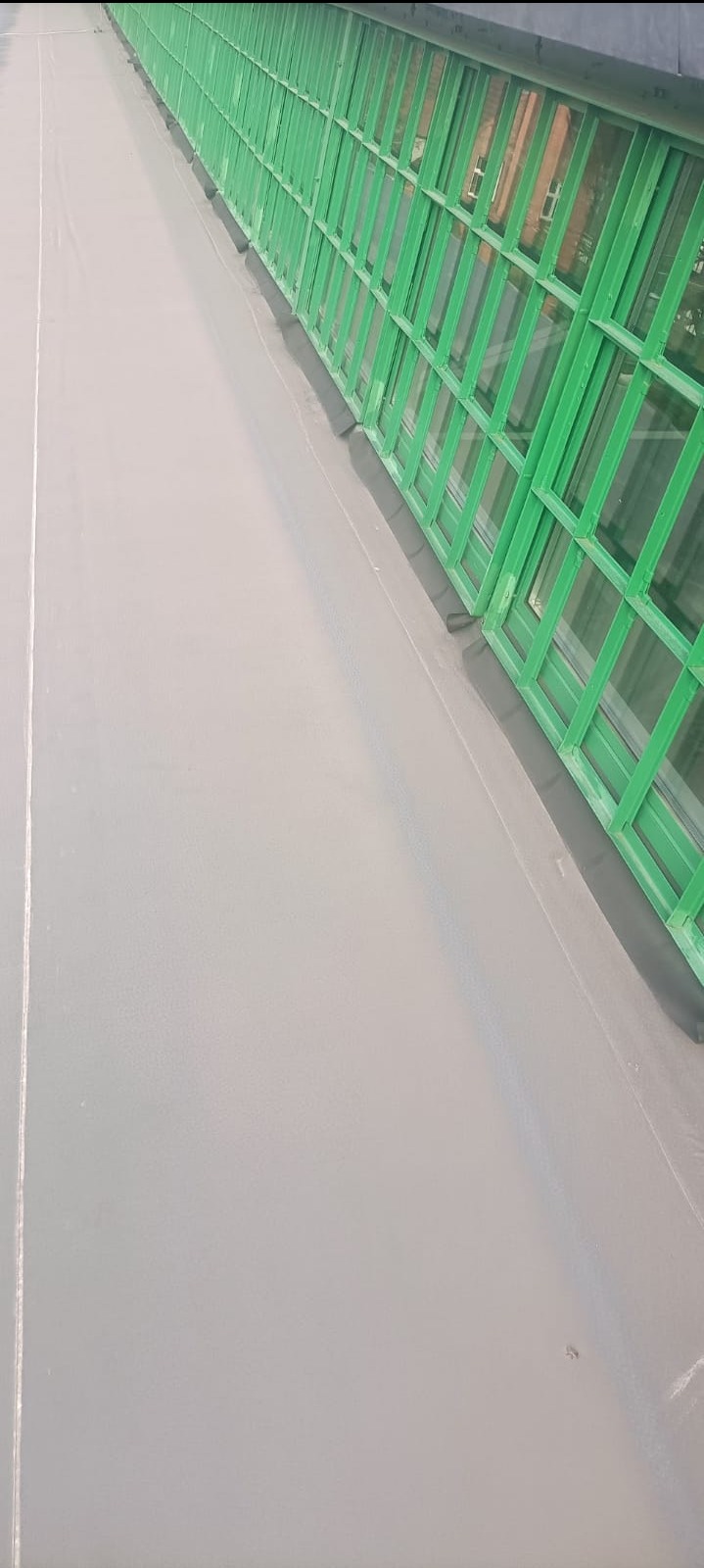 Szare, płaskie pokrycie dachu z widoczną zieloną konstrukcją okienną. Widok z góry, ukośny kadr. Gładka powierzchnia dachu, równomierne wykończenie, odbicia światła.