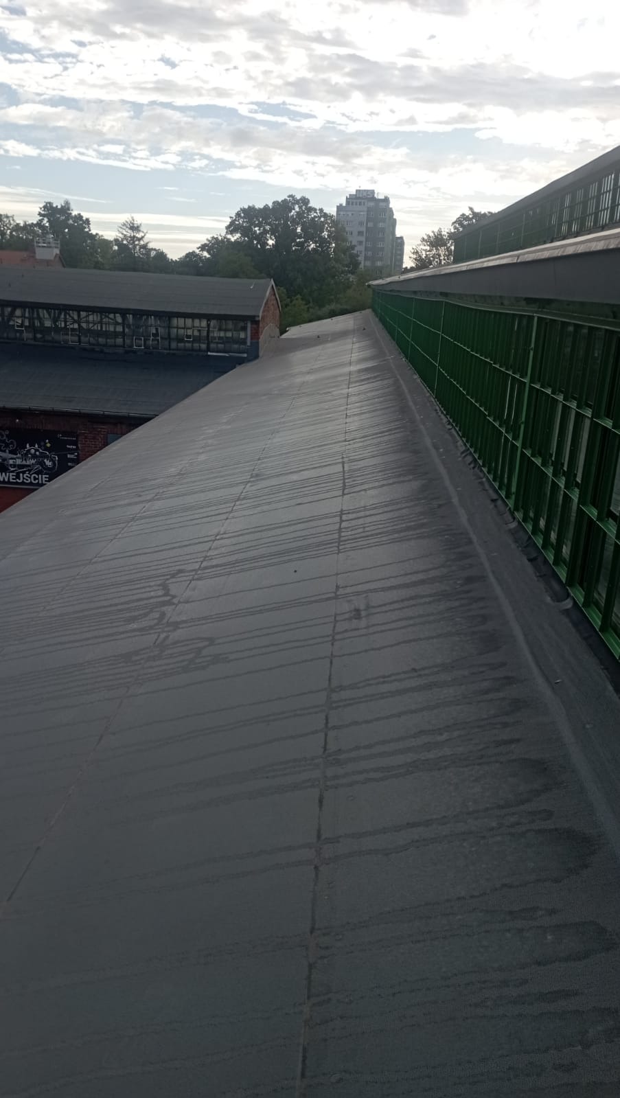 Szary, płaski dach budynku z zieloną metalową barierką. W tle drzewa i budynek mieszkalny. Widoczne ślady po deszczu na powierzchni dachu.