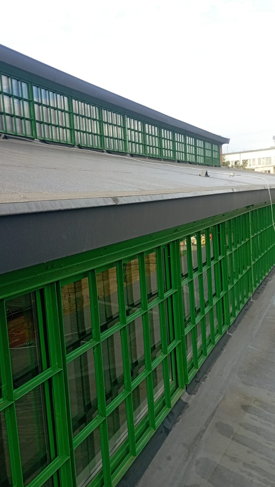 Szary, płaski dach z zielonymi, metalowymi oknami wzdłuż krawędzi. Widok z dołu ukazuje strukturę dachu i regularny układ okien.