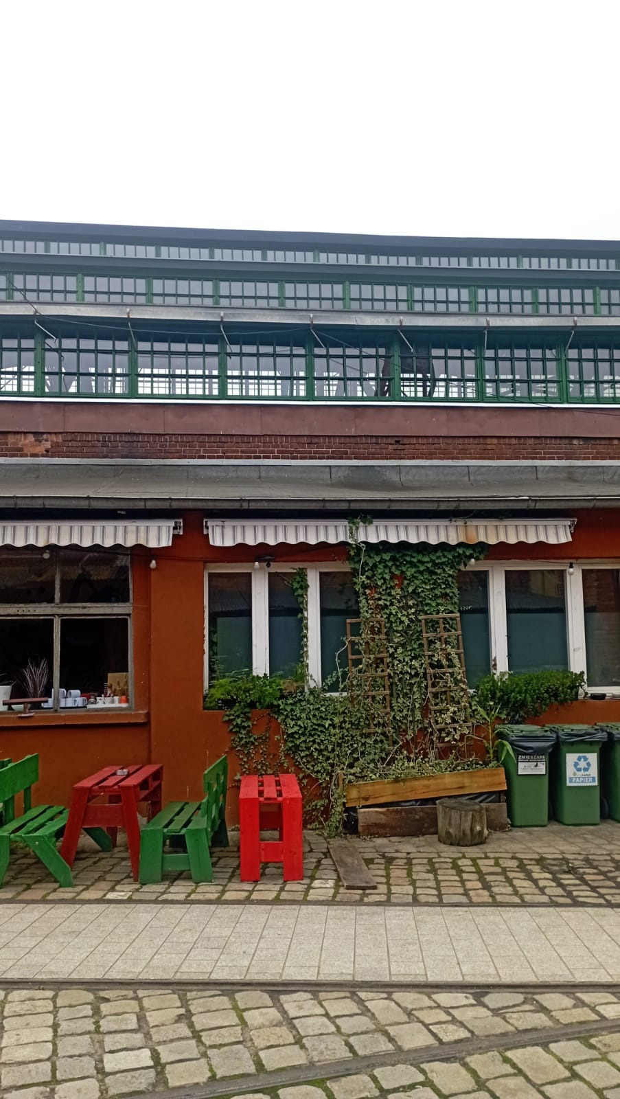Dwupiętrowy budynek z cegły z oknami, zielonymi akcentami i markizami. Na dole kolorowe meble z palet i roślinność, a także kosze na śmieci. Brukowana ścieżka.