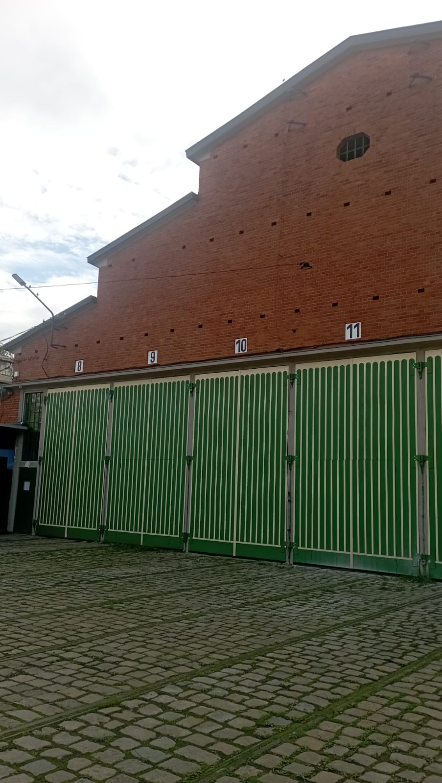 Ceglany budynek z czterema zielonymi bramami z białymi pasami i numerami 8, 9, 10, 11. Brukowana droga przed budynkiem.