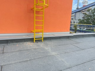 Dach pokryty papą z żółtą drabinką przymocowaną do pomarańczowej ściany budynku przemysłowego. Widok z góry na płaską powierzchnię dachu.