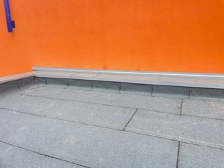 Szare, płaskie pokrycie dachu z papy na tle pomarańczowej ściany. Widoczne detale łączeń i obróbek blacharskich. Minimalistyczne ujęcie z bliska.