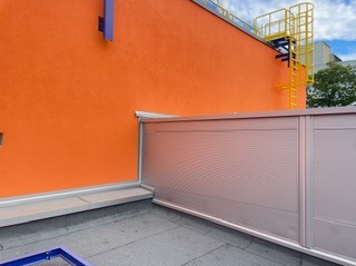 Nowoczesne, metalowe obrzeże dachu na tle pomarańczowej ściany budynku z żółtą drabiną ewakuacyjną. Widok z góry na fragment dachu.