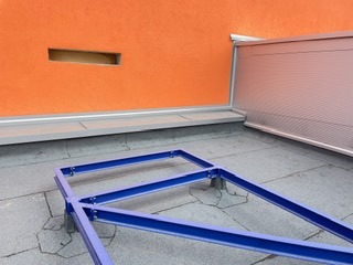 Płaski dach pokryty czarną papą, z widoczną niebieską konstrukcją wsporczą i pomarańczową ścianą z otworem wentylacyjnym w tle. Detal izolacji.