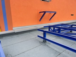 Szare pokrycie dachowe z widoczną konstrukcją wsporczą w kolorze niebieskim na tle pomarańczowej ściany budynku. Widok z góry, oświetlenie dzienne.