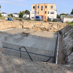 LUBOML.PL DMYTRO BOHOMAZ - Wykop pod budynek z wylaną płytą fundamentową, widoczne zbrojenie i szalunki. W tle budynki mieszkalne i sprzęt budowlany. Ujęcie z góry.