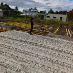 Pracownik w kasku pozdrawia na tle zbrojenia fundament&oacute;w i szalunk&oacute;w. Widoczne niebo z chmurami i budynki w oddali. Układ zbrojenia w regularnych odstępach.