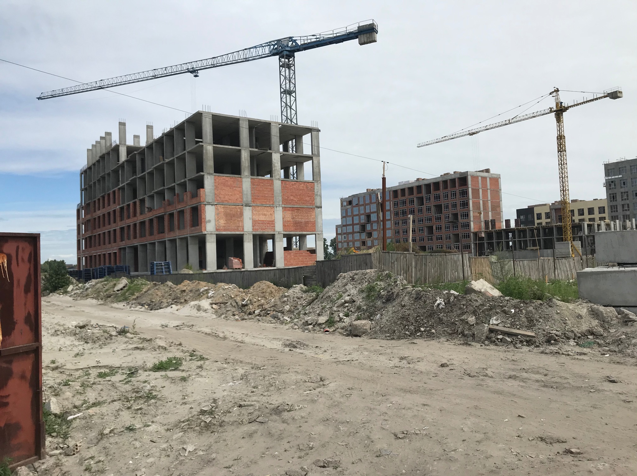 Budowa wielopiętrowego budynku z żelbetową konstrukcją, widoczne ceglane wypełnienia i dwa dźwigi budowlane na tle pochmurnego nieba.