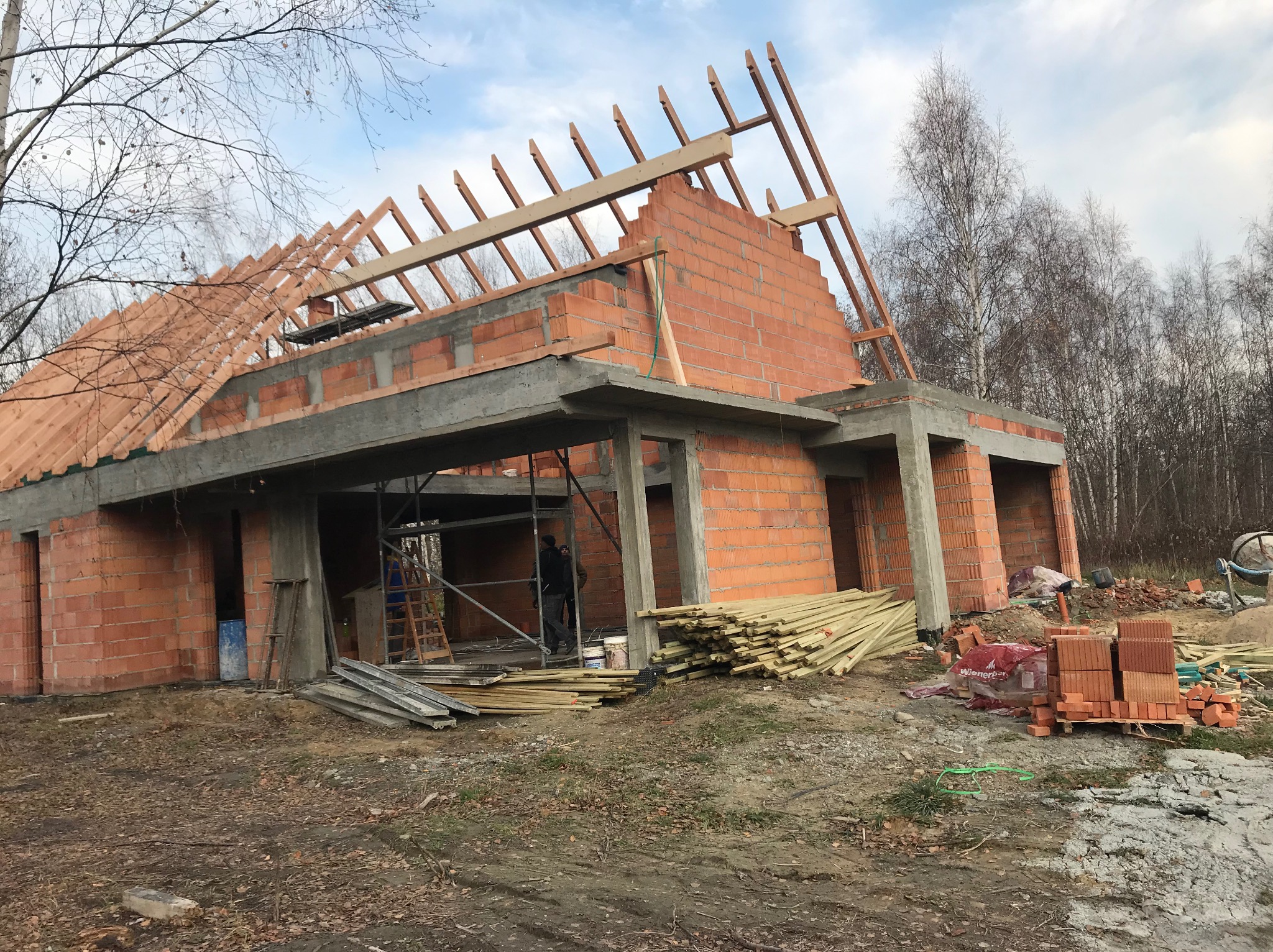 Wznoszenie murów z czerwonej cegły na budowie domu jednorodzinnego. Widoczne elementy konstrukcji dachu i betonowe nadproża. W tle drzewa i niebo.