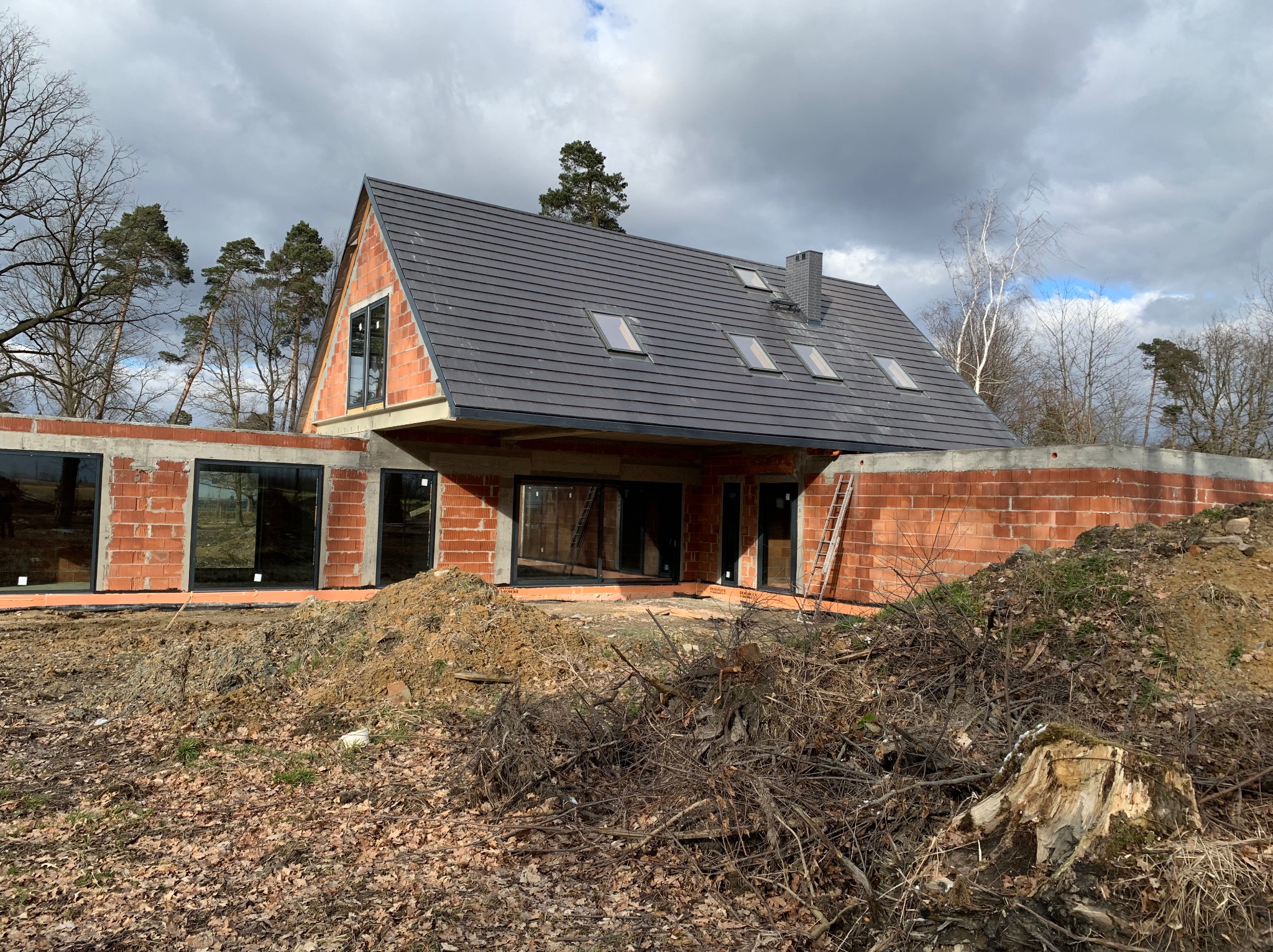 Budowa domu jednorodzinnego w trakcie realizacji: ściany z czerwonej cegły, szary dach z oknami dachowymi, widoczne drzewa w tle i sterta gałęzi na pierwszym planie.