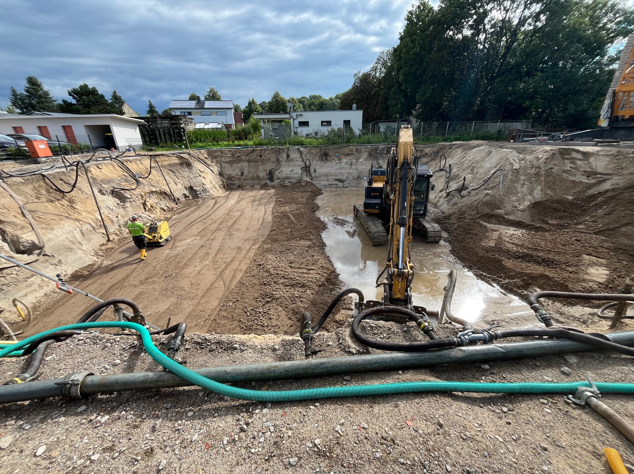 Wykop pod fundamenty z pracującą koparką i pompami odwadniającymi. Widoczny pracownik obsługujący zagęszczarkę. Pochmurne niebo nad terenem budowy.