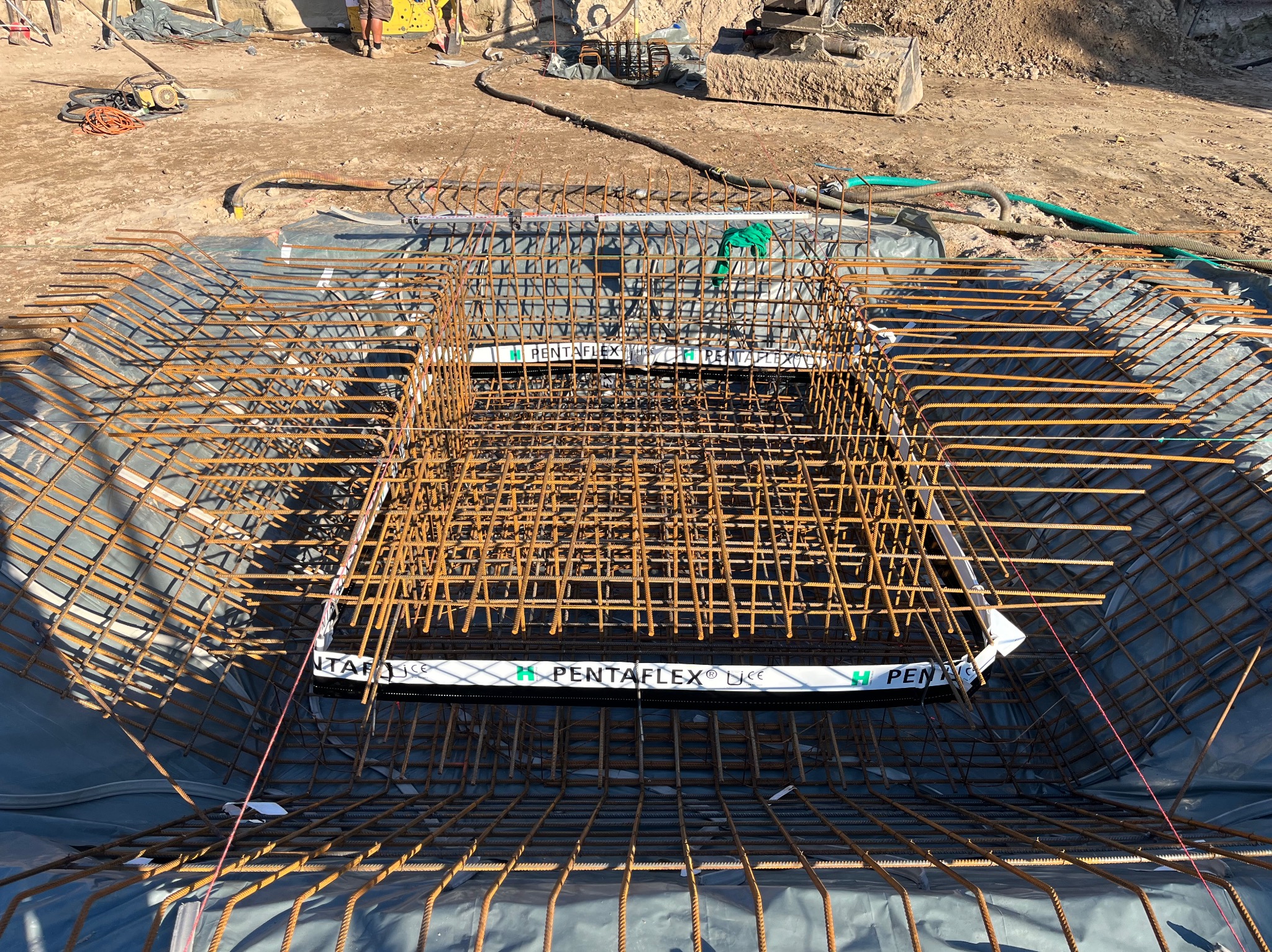 Siatka zbrojeniowa w wykopie zabezpieczonym folią, widok z góry. Na pierwszym planie wystające pręty, w tle ziemia i sprzęt budowlany.