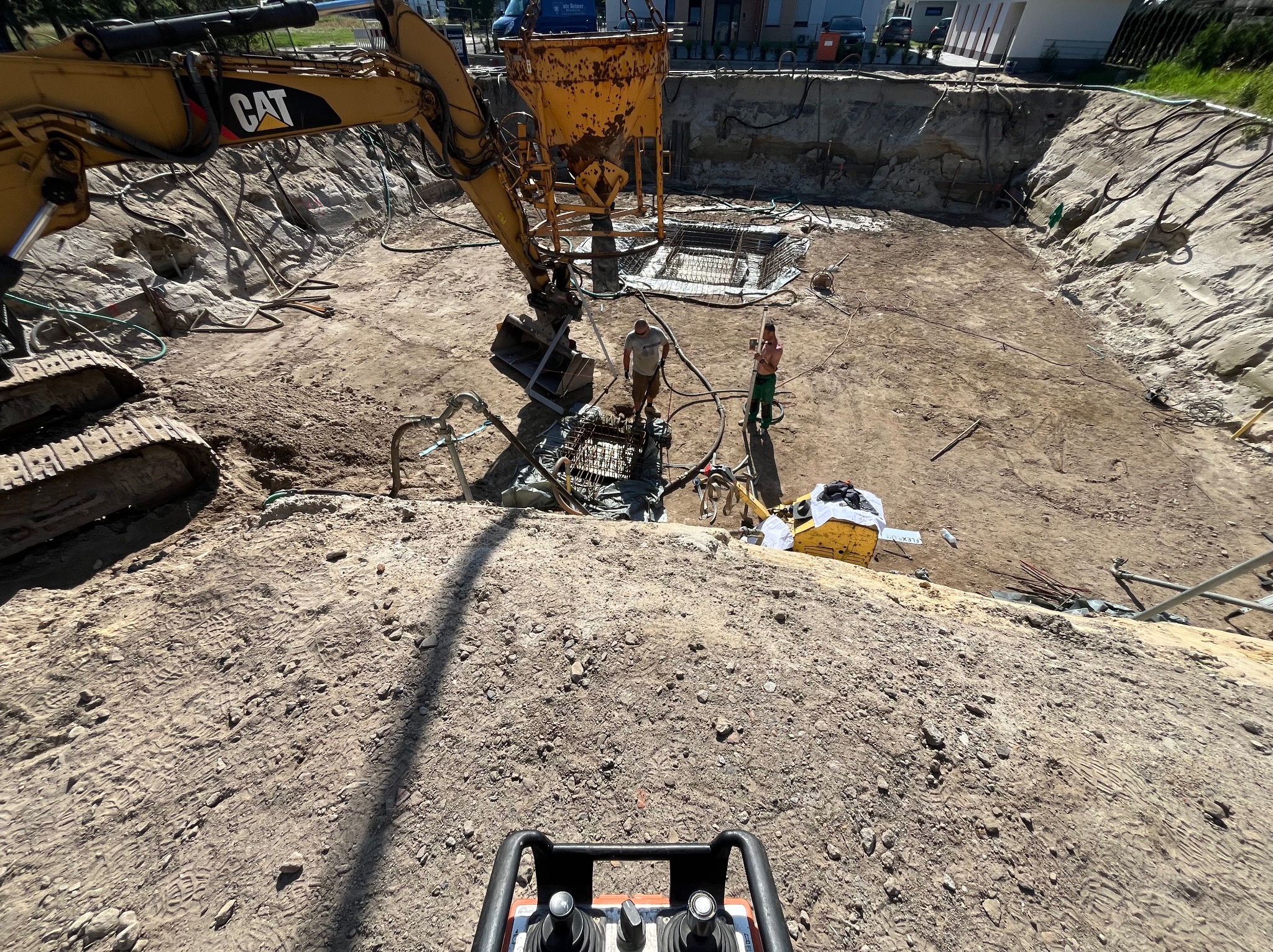 Żółty dźwig CAT opuszcza beton do szalunku zbrojeniowego w wykopie fundamentowym. Dwóch pracowników nadzoruje proces, w tle widoczny teren budowy i budynki mieszkalne.