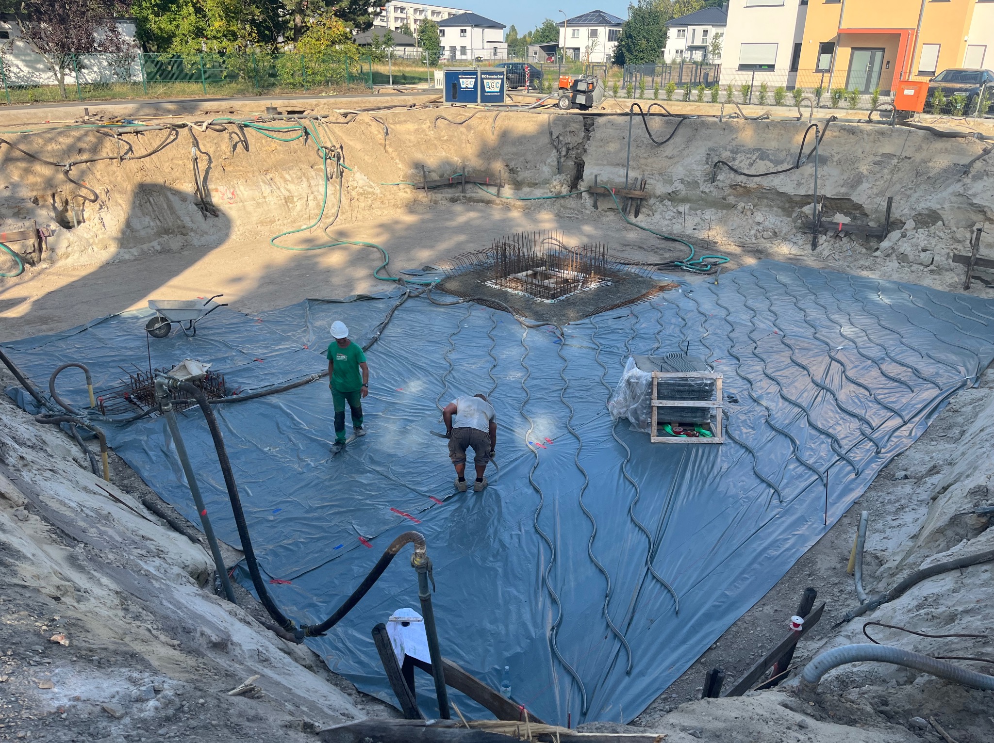 Przygotowanie pod wylewkę fundamentów: wykop, folia izolacyjna, zbrojenie. Dwóch pracowników w trakcie układania instalacji pod wylewkę. Widok z góry na plac budowy.