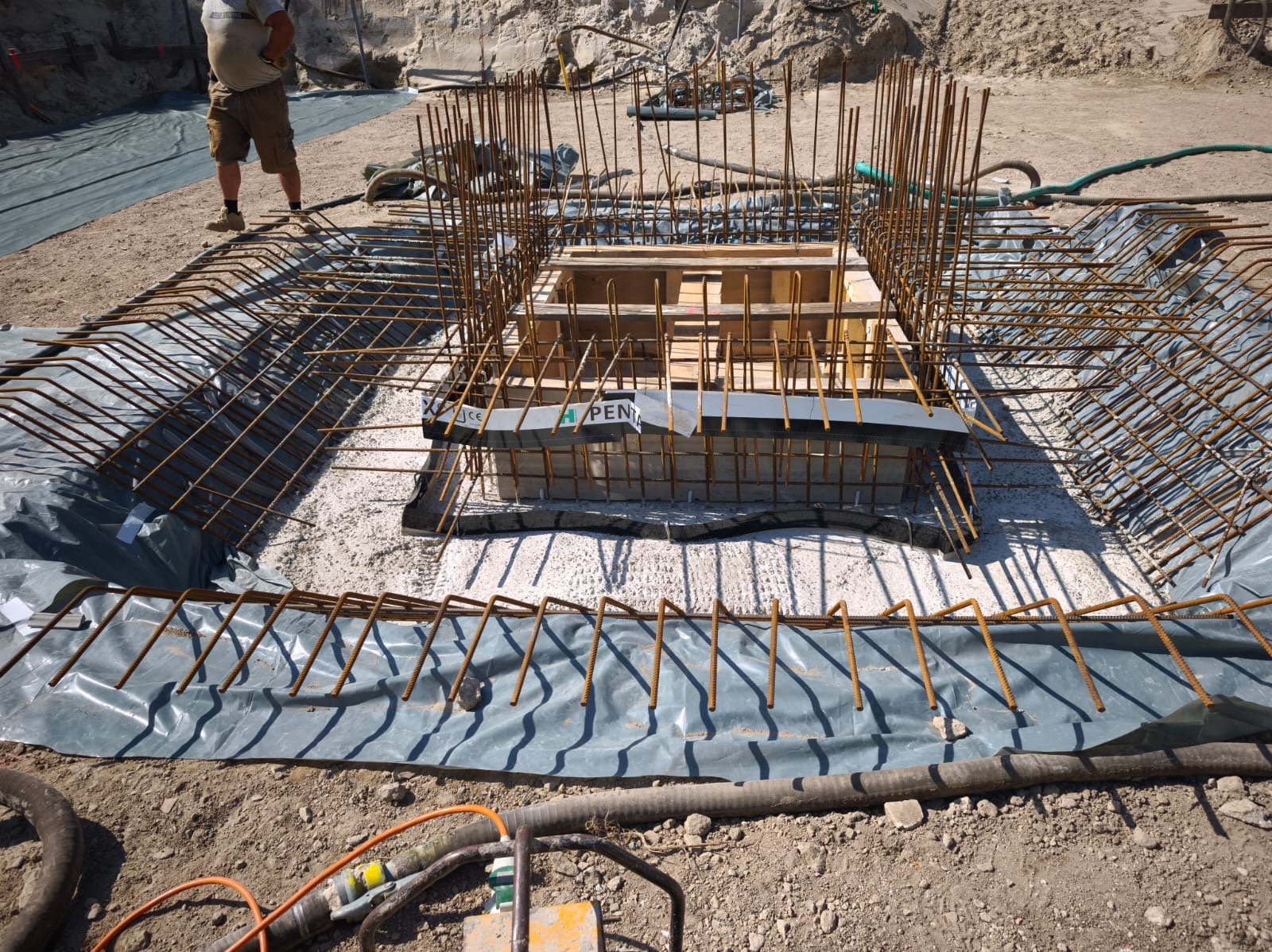 Zbrojenie fundamentu pod budynek, widoczne pręty stalowe i szalunek. Na dnie wykopu folia izolacyjna, częściowo zalane betonem. Ujęcie z góry, widoczny fragment sylwetki człowieka.