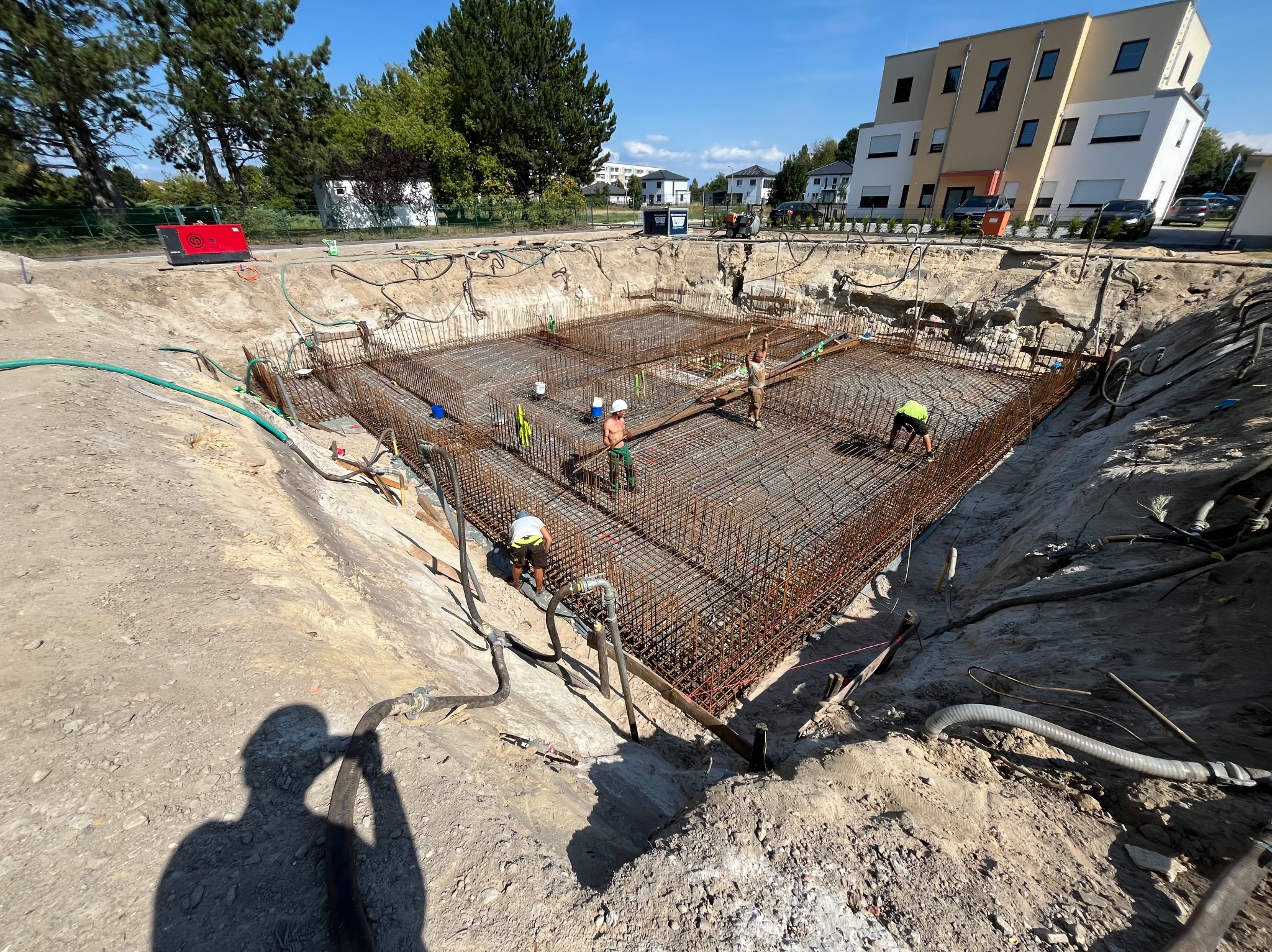 Zbrojenie fundamentów pod budynek mieszkalny. Widoczni pracownicy w kaskach i kamizelkach odblaskowych, zbrojenie z metalowych prętów, wykop ziemny.