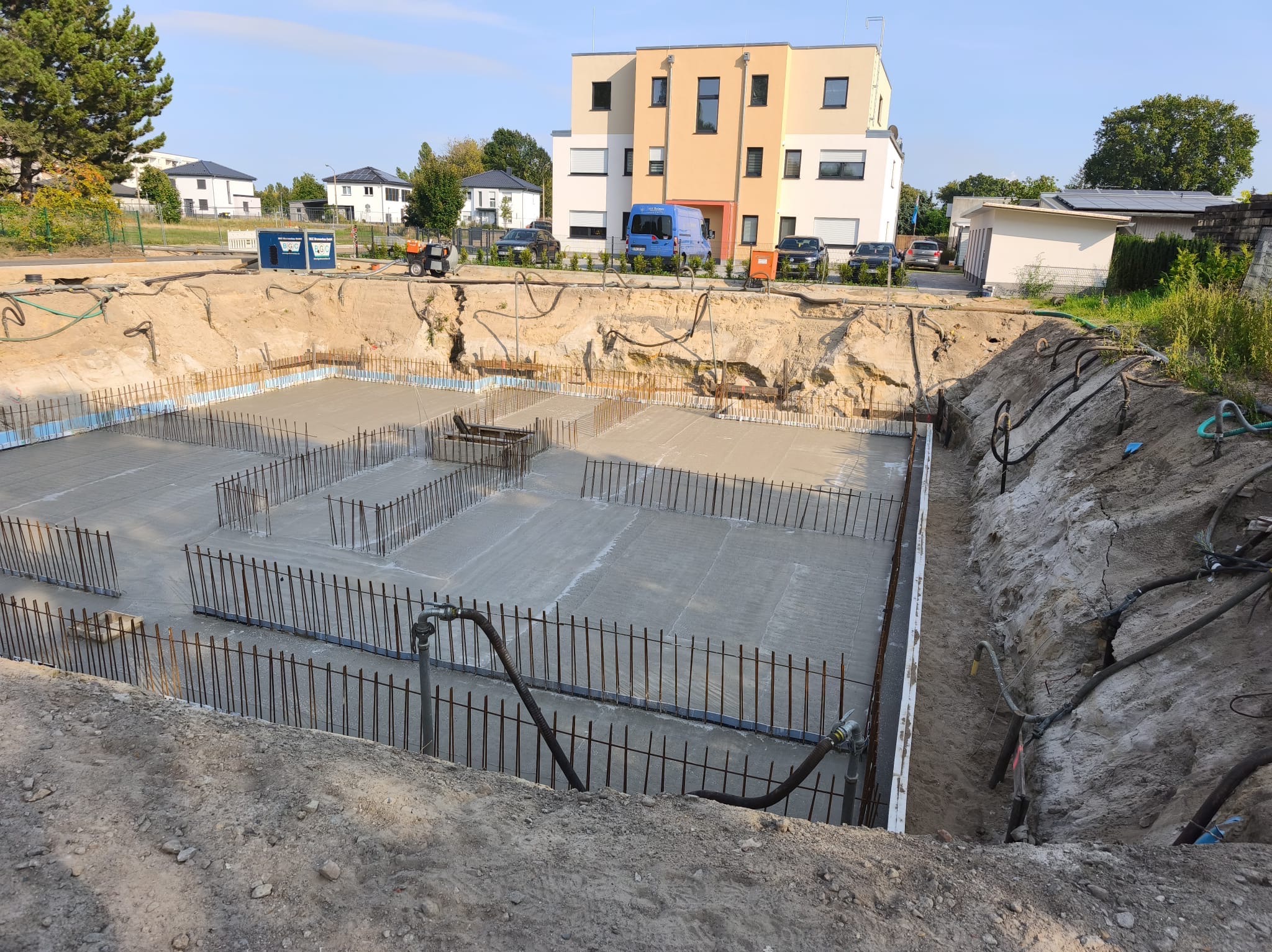Wykop pod budynek z wylaną płytą fundamentową, widoczne zbrojenie i szalunki. W tle budynki mieszkalne i sprzęt budowlany. Ujęcie z góry.