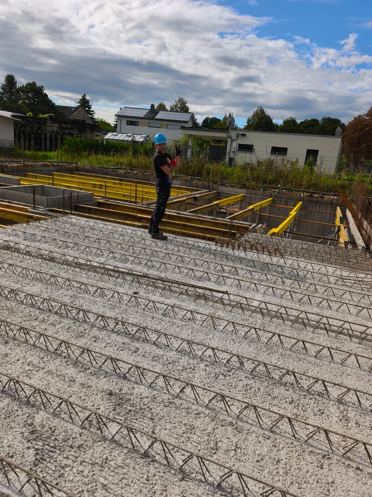 Pracownik w kasku pozdrawia na tle zbrojenia fundamentów i szalunków. Widoczne niebo z chmurami i budynki w oddali. Układ zbrojenia w regularnych odstępach.