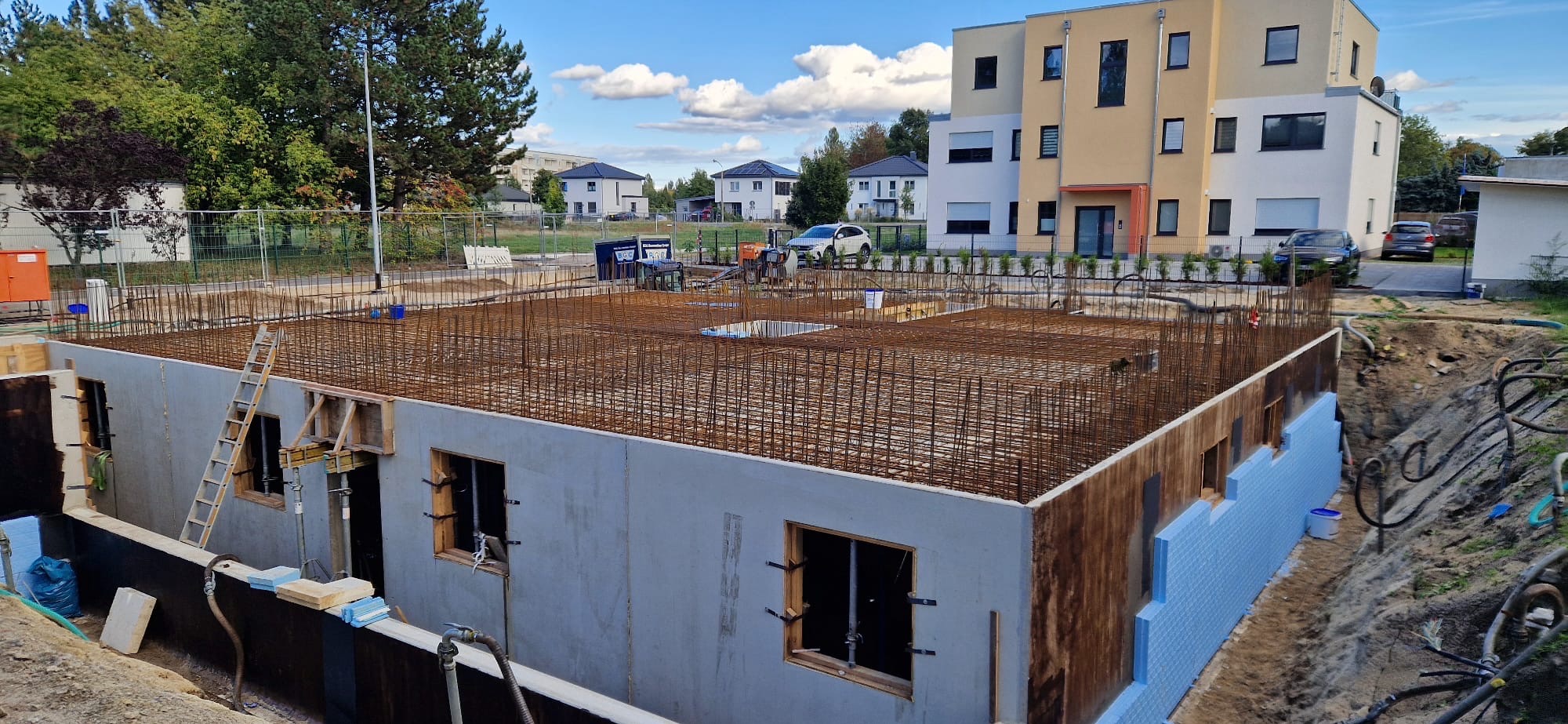 Przygotowany szalunek z oknami i izolacją termiczną, zbrojenie na fundamencie domu jednorodzinnego. Widoczne budynki w tle i niebo.