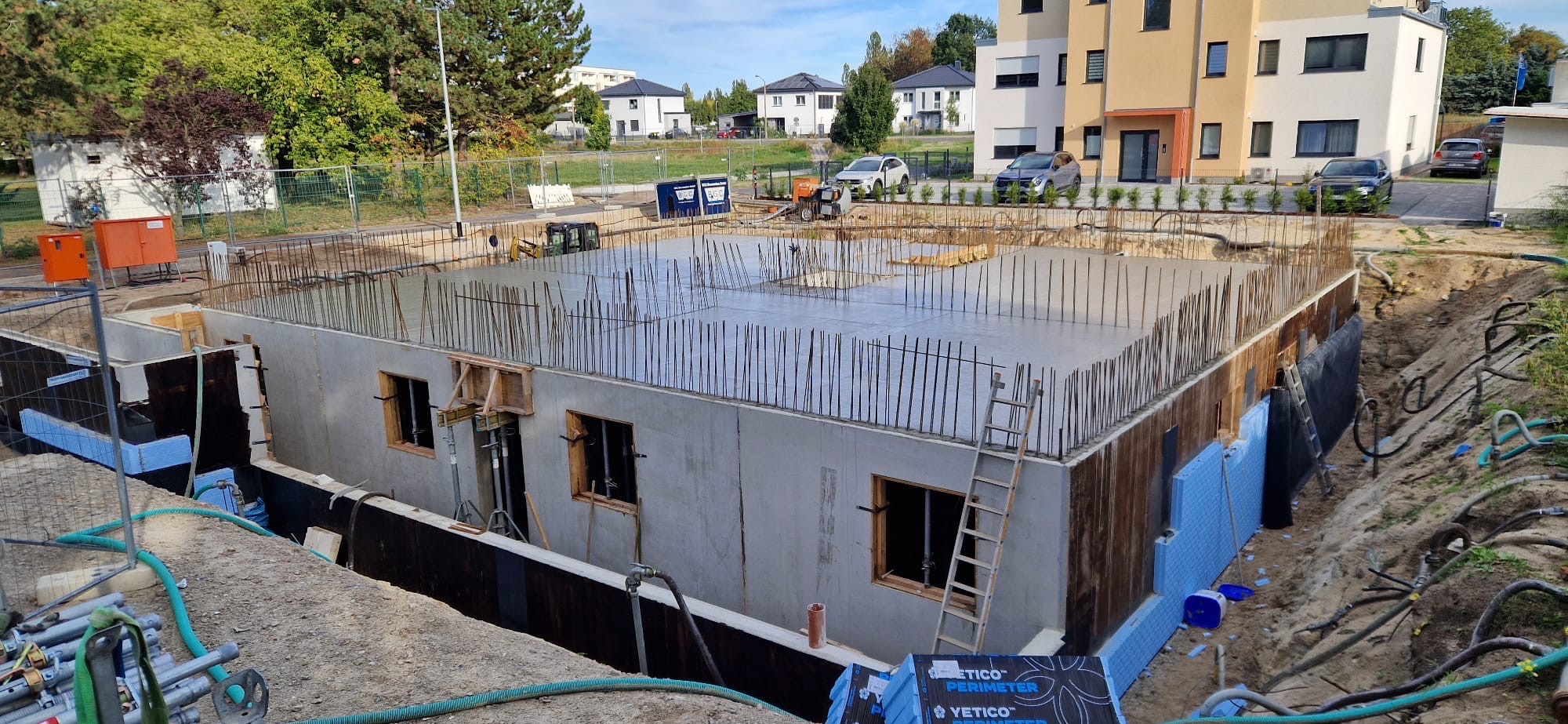 Wylany fundament budynku z wystającymi prętami zbrojeniowymi, widoczne okna piwniczne i ocieplenie. Na zewnątrz budynku sprzęt budowlany i zaparkowane samochody.