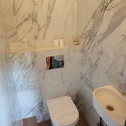 Firma Remontowo-Wykończeniowa T.M.-BUD Tymoteusz Michalski - Elegancka, mała łazienka z marmurowymi płytkami, podwieszaną toaletą i umywalką. Na podłodze drewnopodobne płytki i szary dywanik.