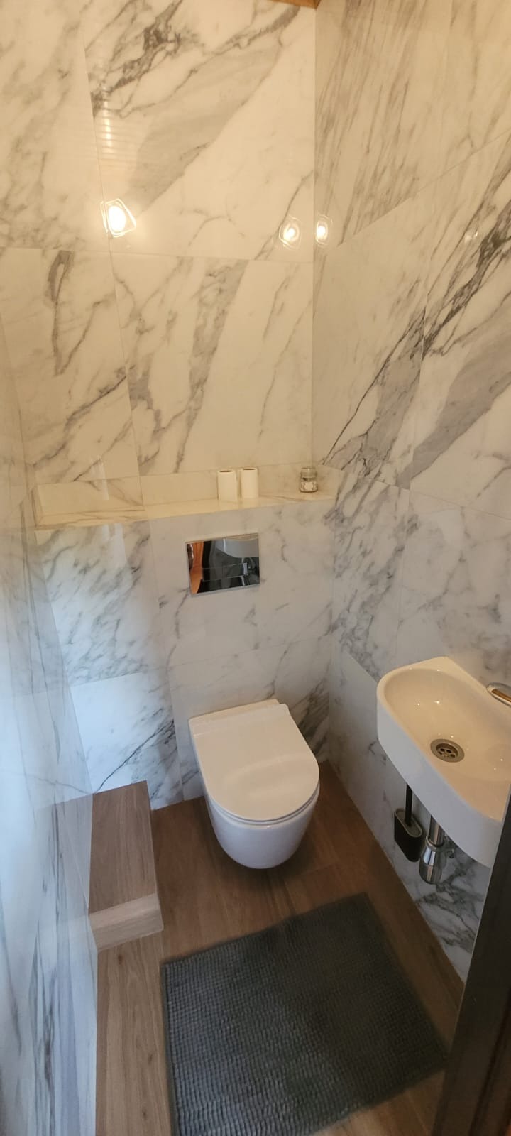 Elegancka, mała łazienka z marmurowymi płytkami, podwieszaną toaletą i umywalką. Na podłodze drewnopodobne płytki i szary dywanik.