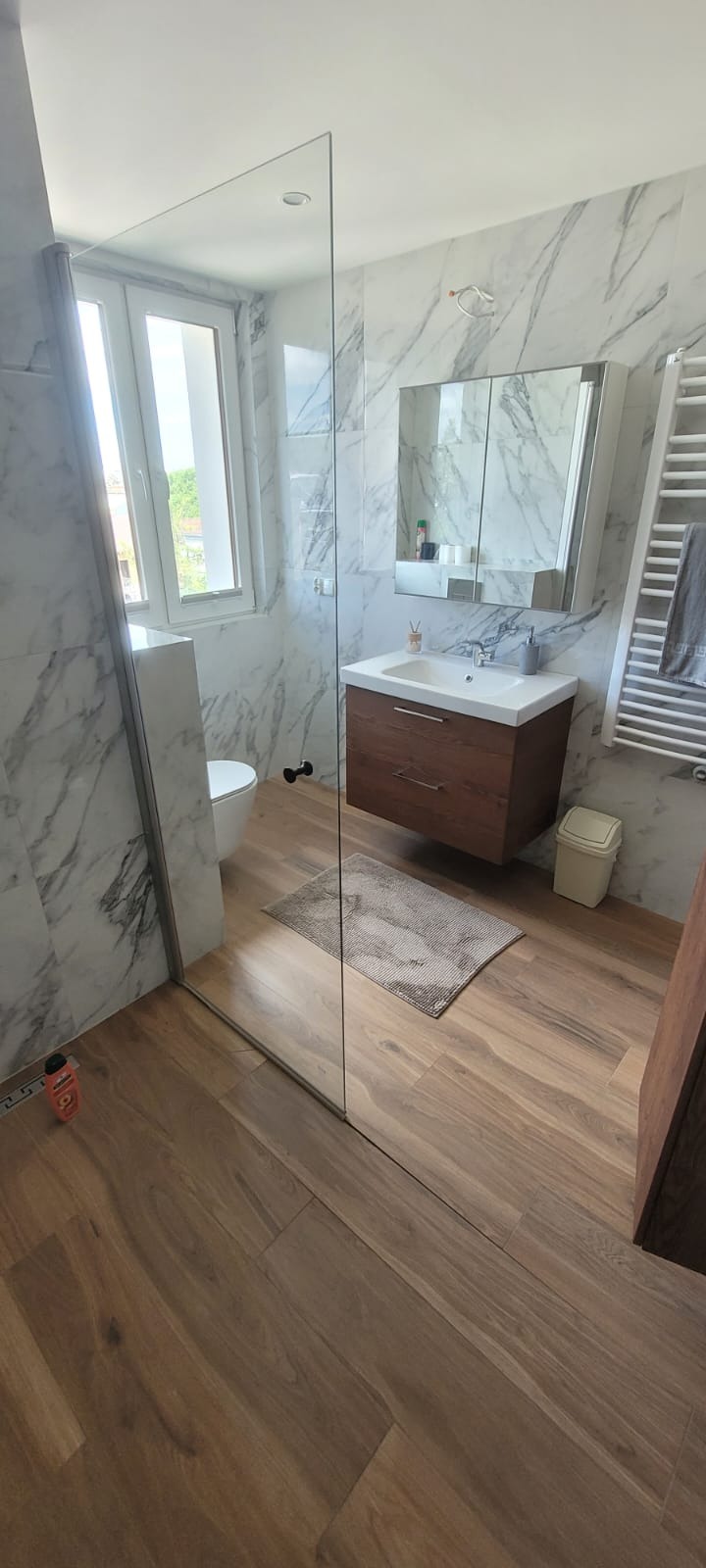Nowoczesna łazienka z kabiną prysznicową typu walk-in, marmurowymi płytkami na ścianach i drewnianą szafką pod umywalką. Widoczne drewniane panele podłogowe.