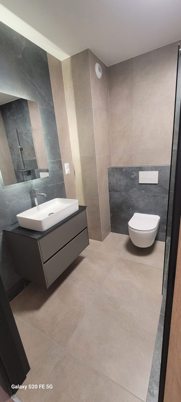 Nowoczesna łazienka po remoncie: podwieszana toaleta, umywalka na szafce z szufladami, szare płytki imitujące kamień i beżowe płytki podłogowe.