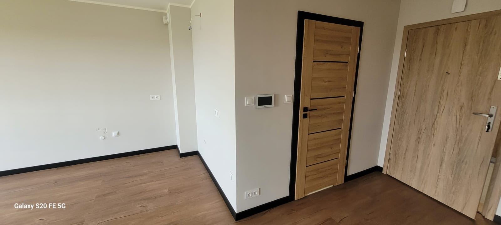 Nowoczesne wnętrze: dwie drewniane drzwi z czarnymi ościeżnicami, panele podłogowe i ściany w odcieniach szarości. Widoczne gniazdka elektryczne i panel sterowania.