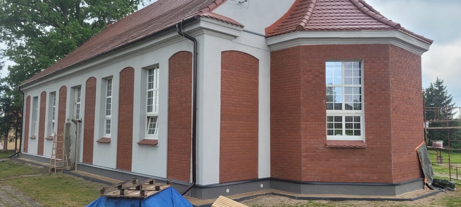 Odnowiona elewacja budynku z ceglanymi wstawkami i białymi kolumnami okiennymi. Widoczne okna z białymi ramami i brązowy dach z dachówki.