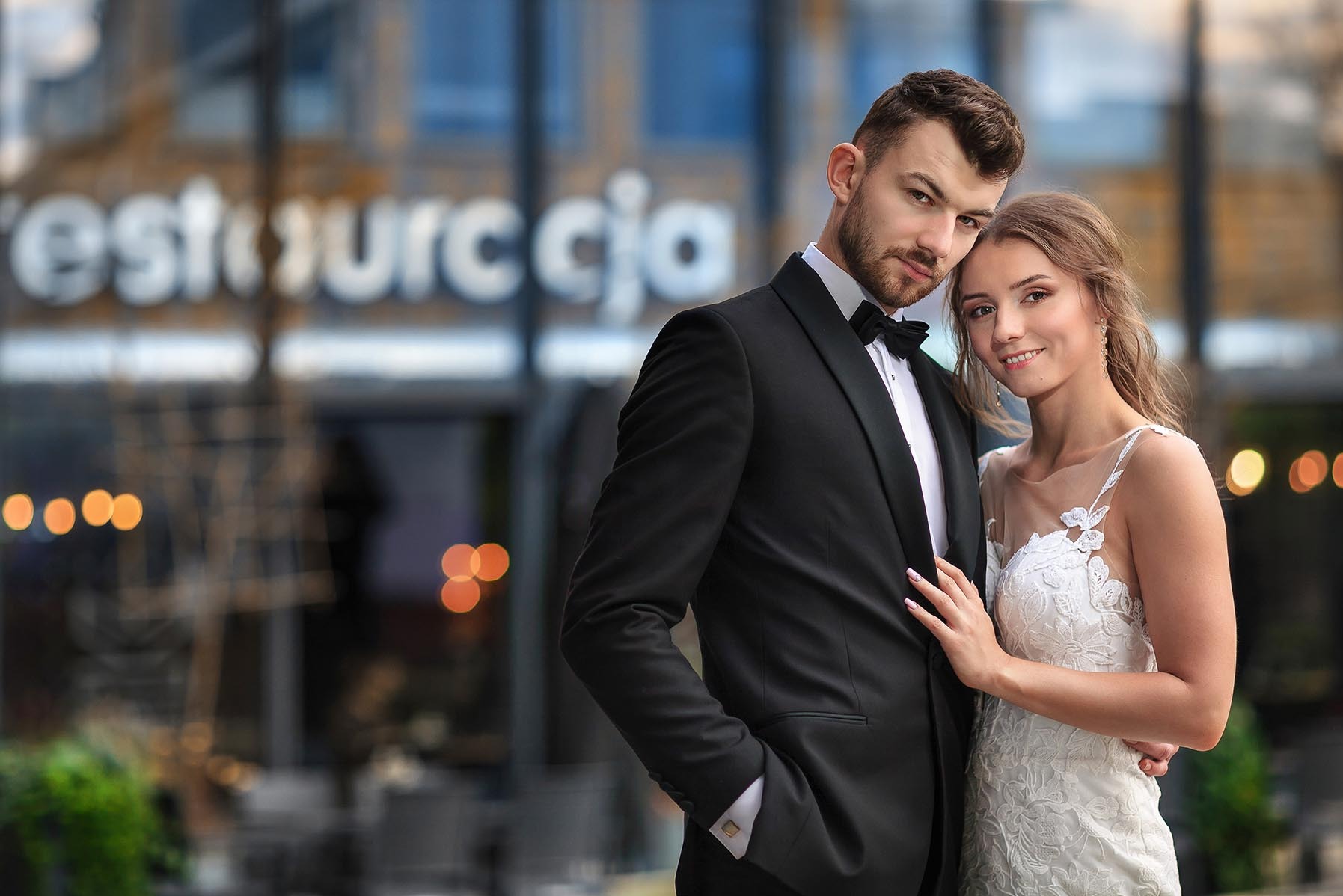 Elegancka para młoda w objęciach na tle restauracji z rozmytymi światłami, on w smokingu, ona w koronkowej sukni, subtelny uśmiech, intymna atmosfera.