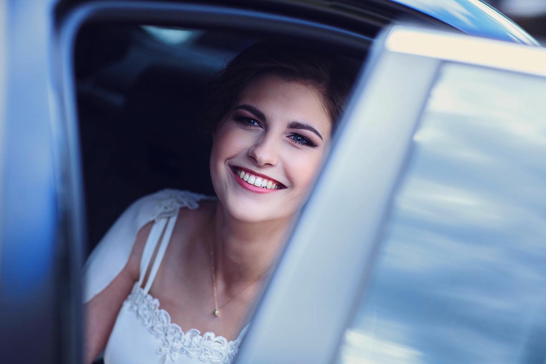Uśmiechnięta panna młoda w białej sukni, spoglądająca zza otwartych drzwi samochodu. Widoczny makijaż i delikatna biżuteria. Subtelne oświetlenie.