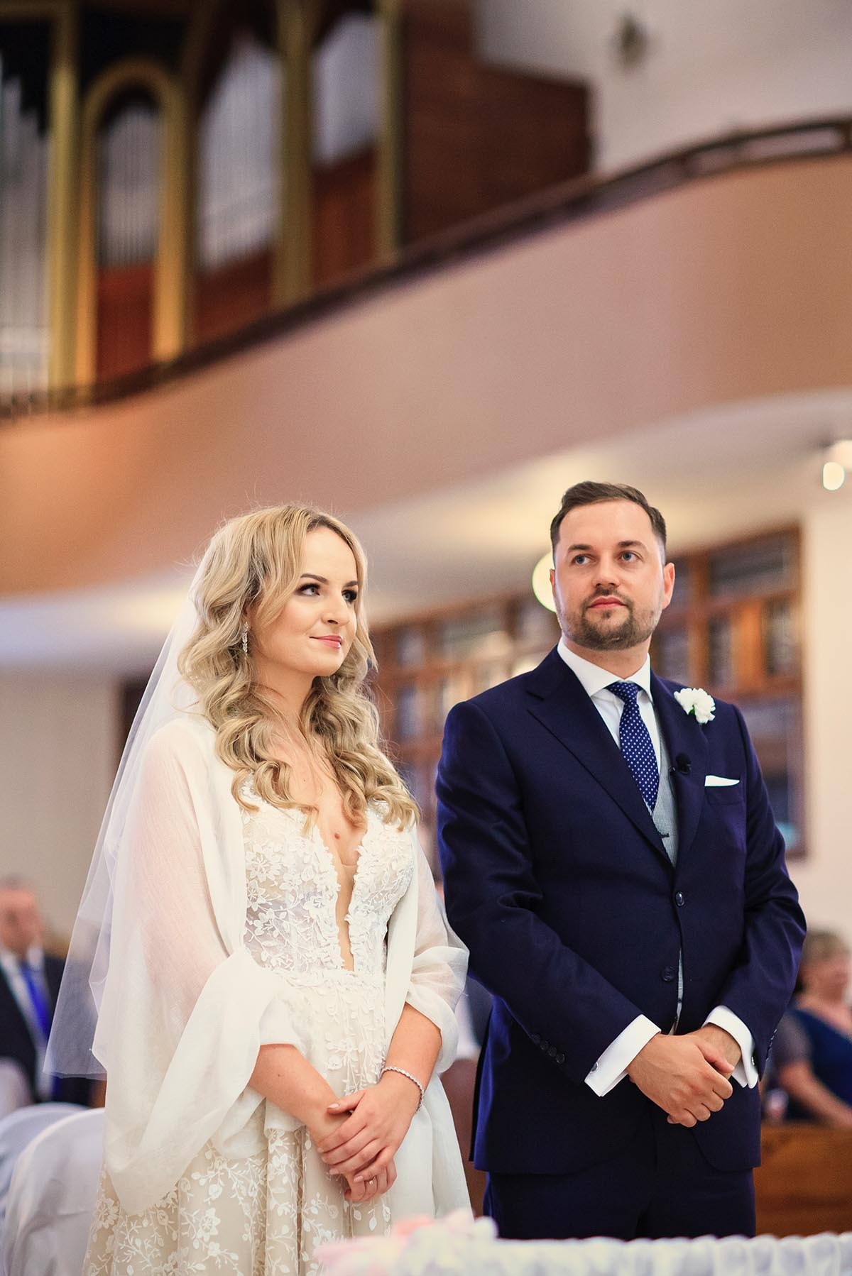 Para młoda podczas ceremonii ślubnej w kościele. Panna młoda w koronkowej sukni i welonie, pan młody w granatowym garniturze. Dekoracje kwiatowe w tle.