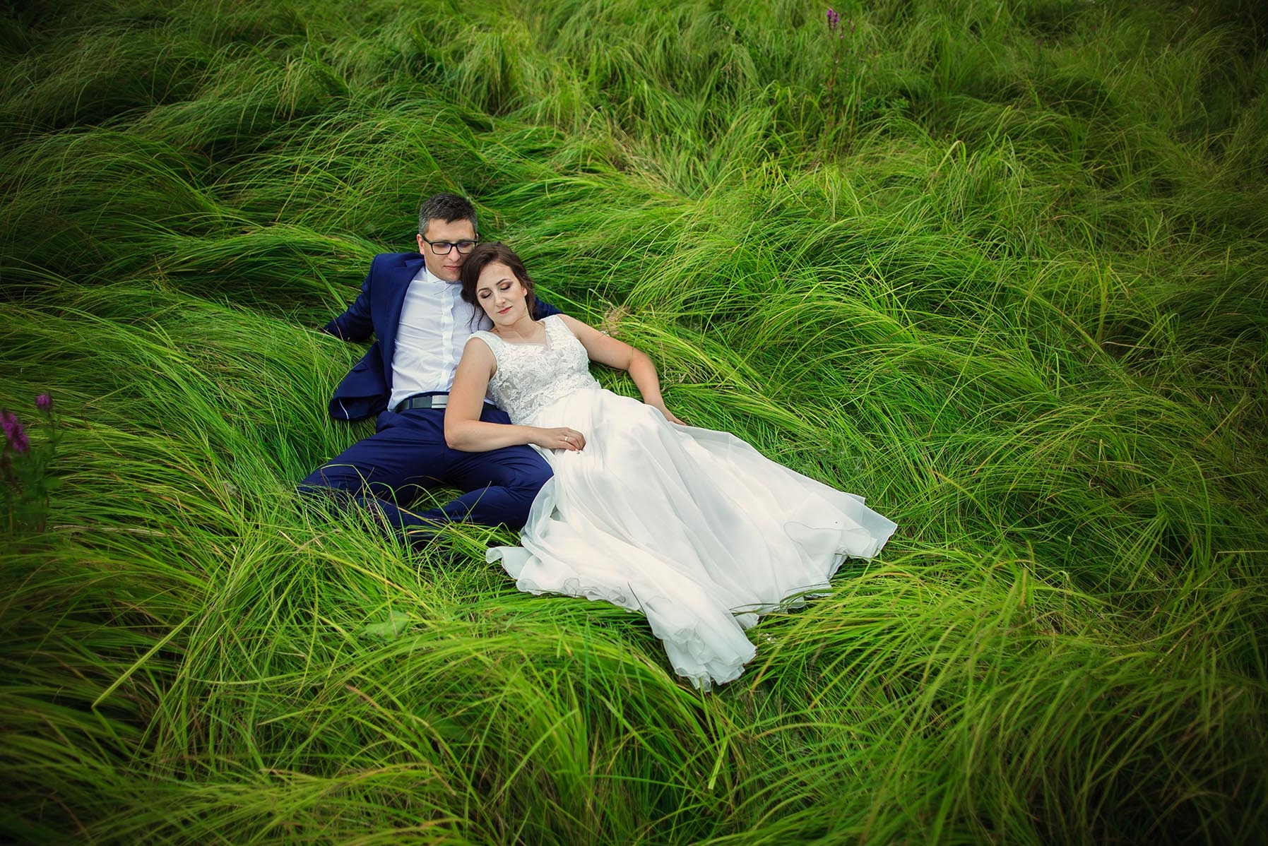 Para młoda leży na łące w wysokiej trawie. Panna młoda w białej sukni, pan młody w granatowym garniturze. Romantyczna sesja w plenerze, naturalne światło.
