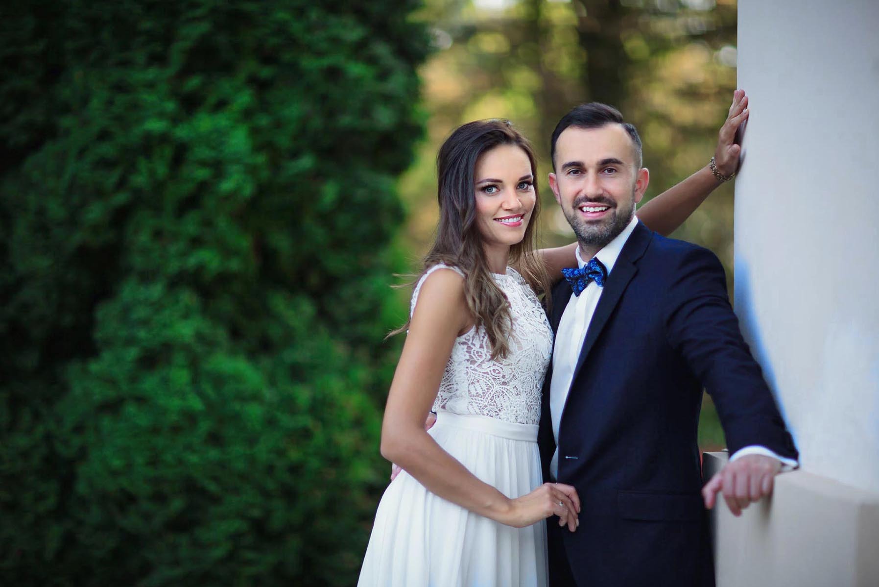 Uśmiechnięta para młoda w eleganckich strojach pozuje na tle zieleni i białej ściany. Pan w granatowym garniturze i muszce, pani w koronkowej sukni.
