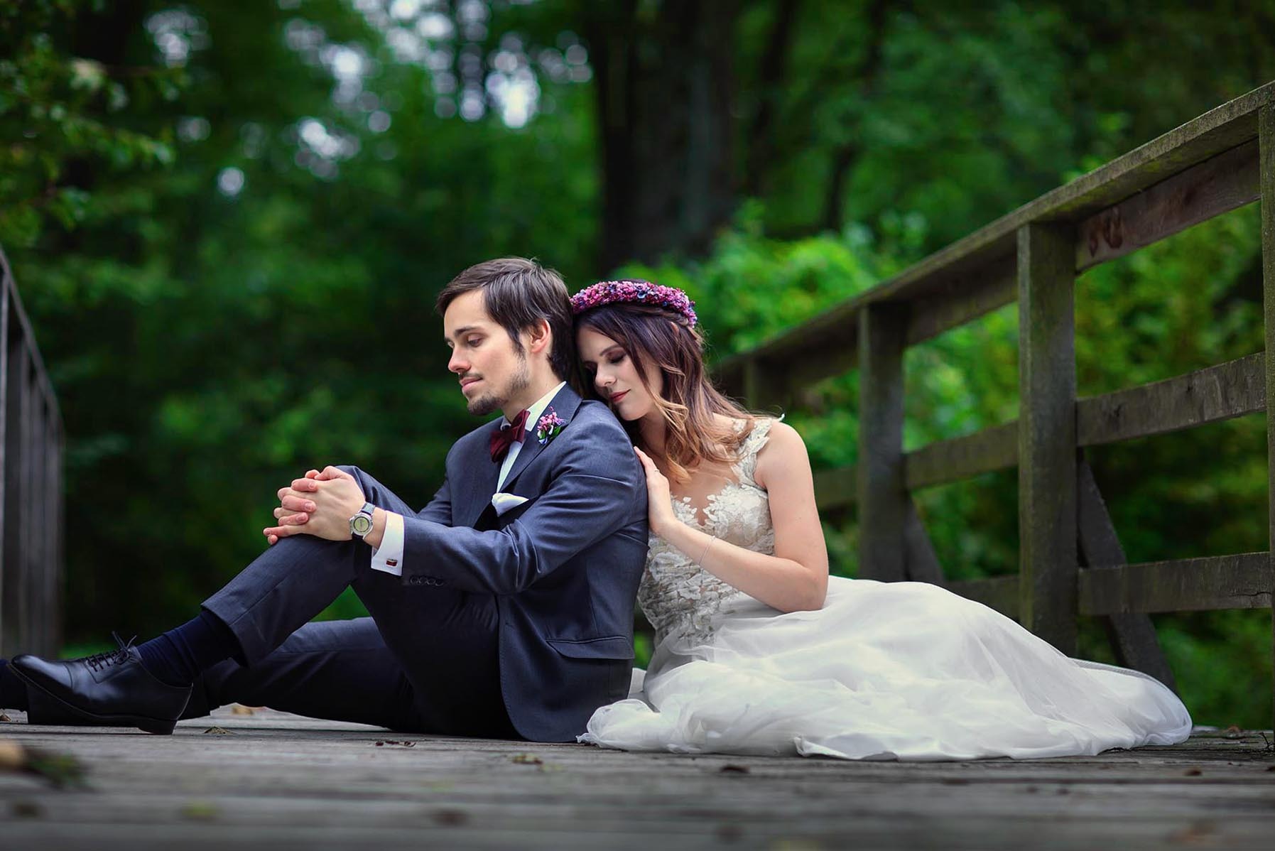 Para młoda siedzi na drewnianym moście w lesie. Panna młoda w wianku opiera głowę na ramieniu pana młodego w granatowym garniturze i bordowej muszce.