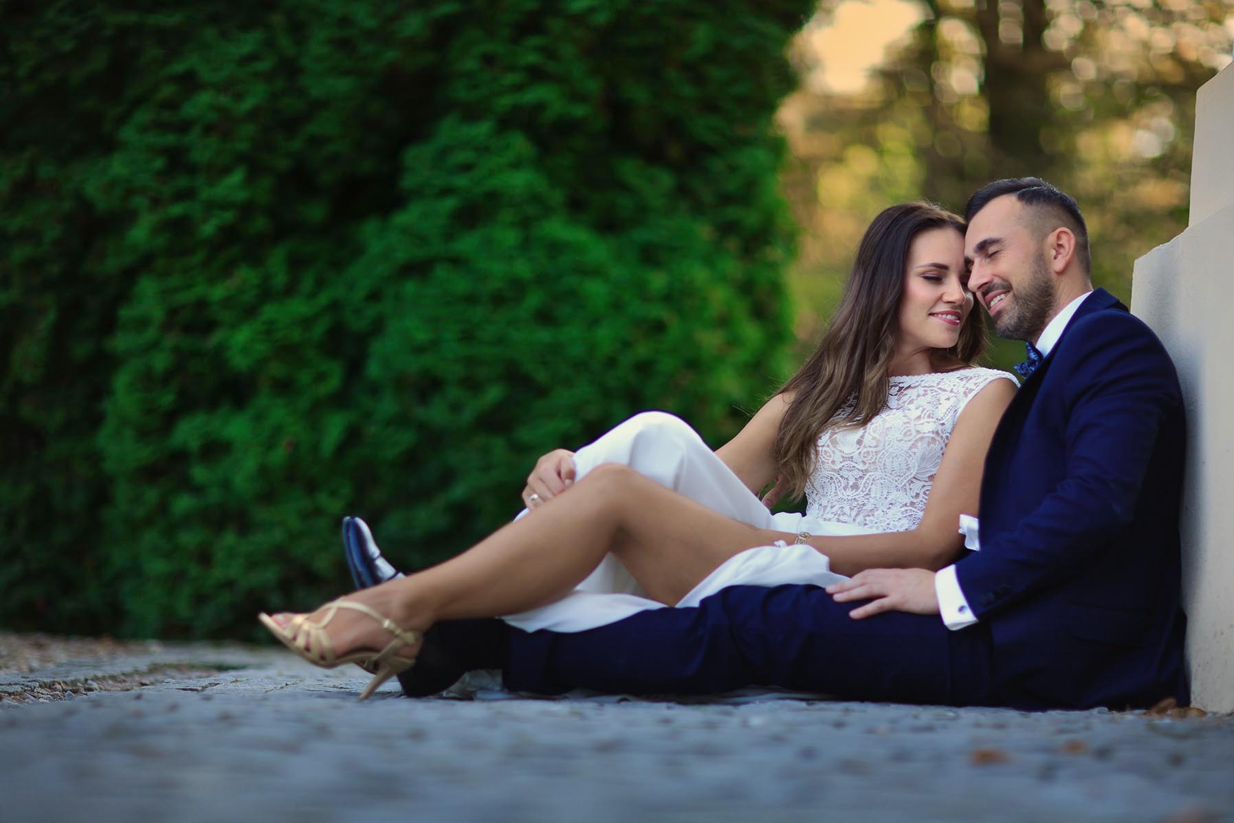 Para młoda siedzi na ziemi, opierając się o ścianę, w tle zieleń. Ona w białej sukni z koronkową górą, on w granatowym garniturze z muszką.