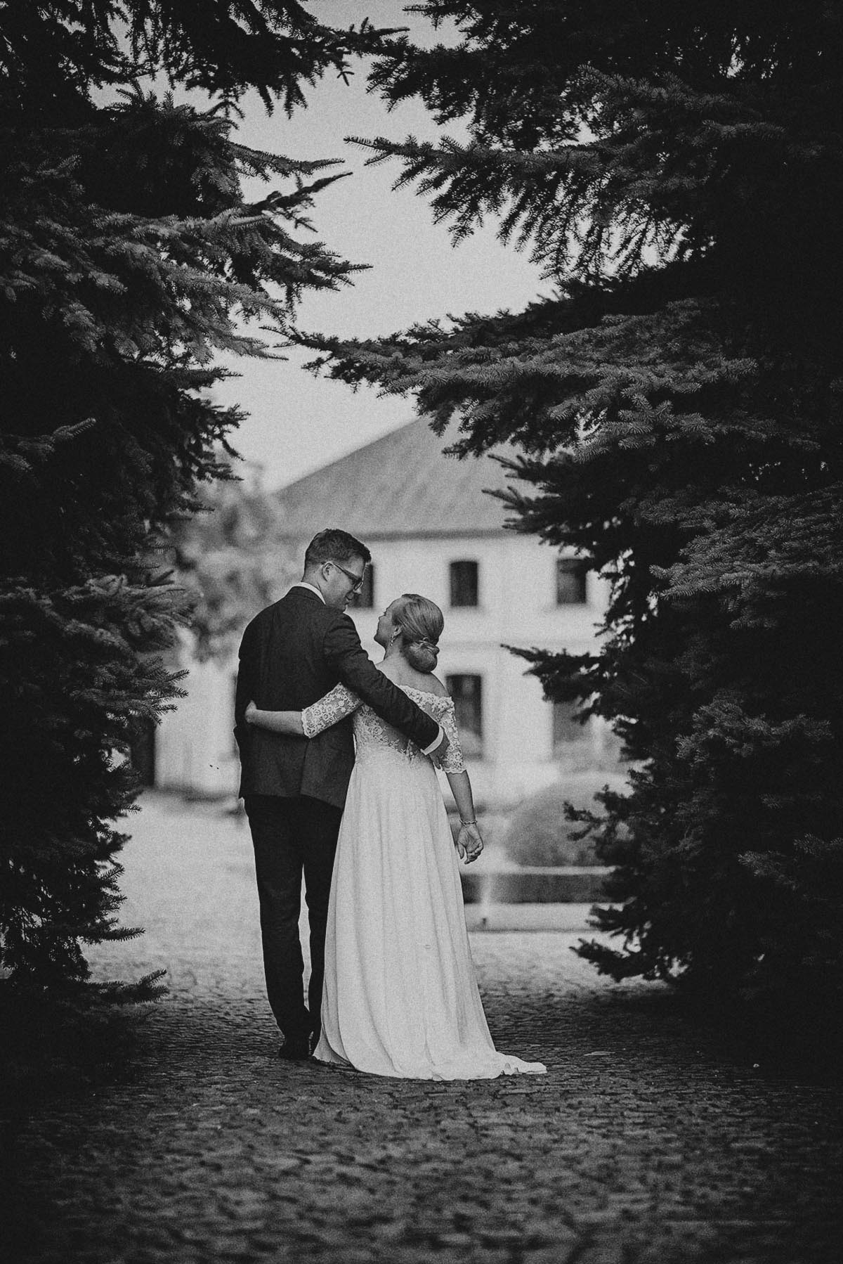 Czarno-białe zdjęcie pary młodej, obejmującej się na tle budynku, otoczonej drzewami iglastymi. Panna młoda w sukni z koronkowymi rękawami, pan młody w garniturze.