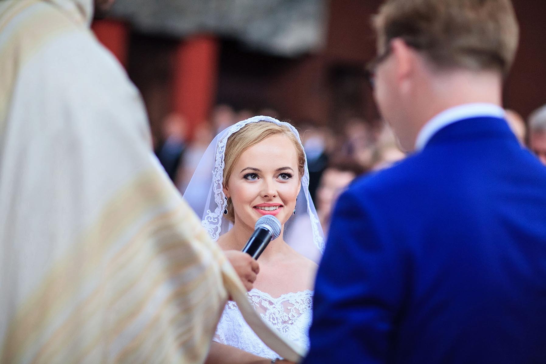 Panna młoda z mikrofonem podczas ceremonii ślubnej w kościele, otoczona przez księdza i pana młodego. Uśmiechnięta kobieta w welonie i koronkowej sukni.