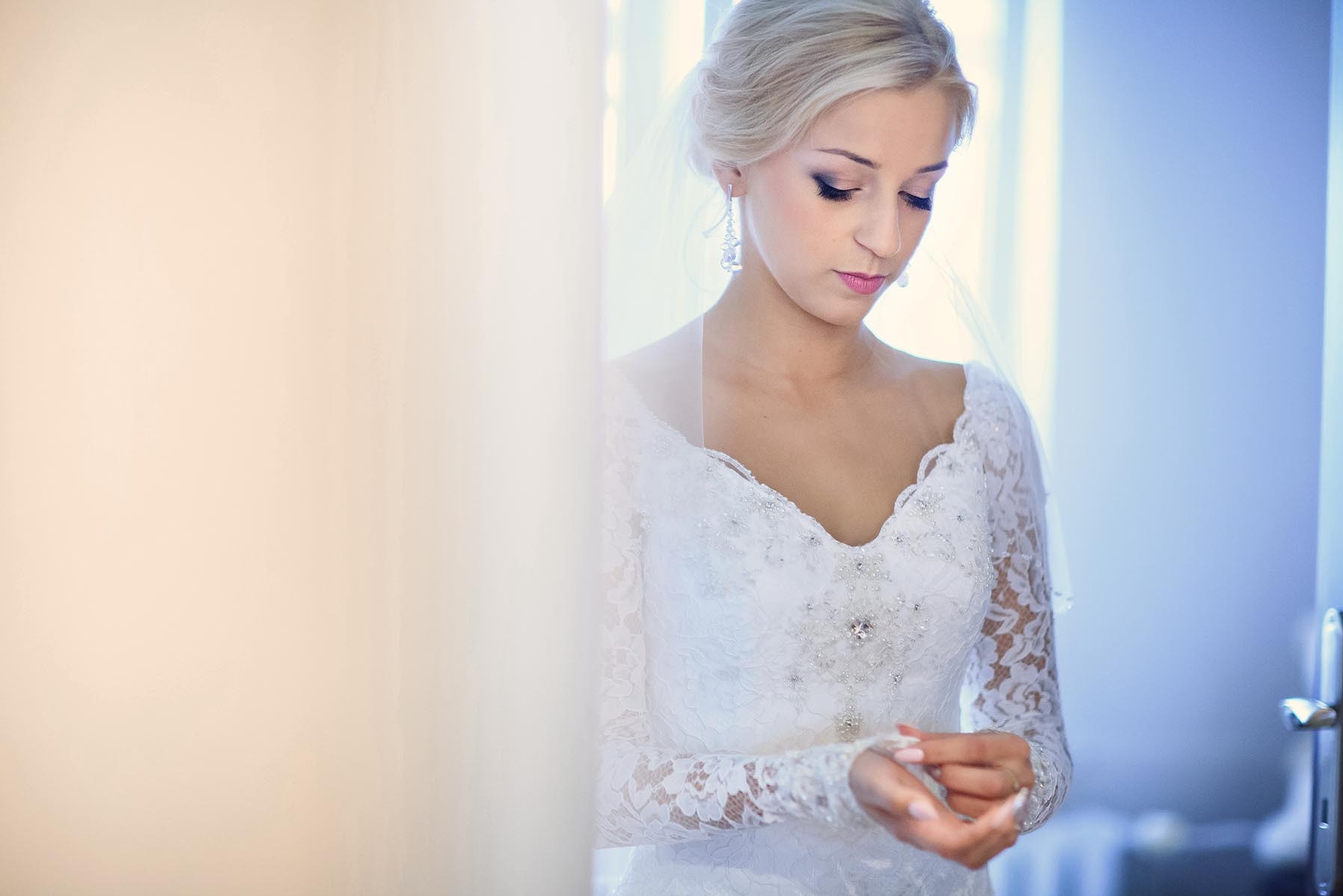 Panna młoda w koronkowej sukni z długim rękawem, z delikatnym makijażem, spogląda w dół, poprawiając mankiet. W tle subtelne światło i fragment drzwi.