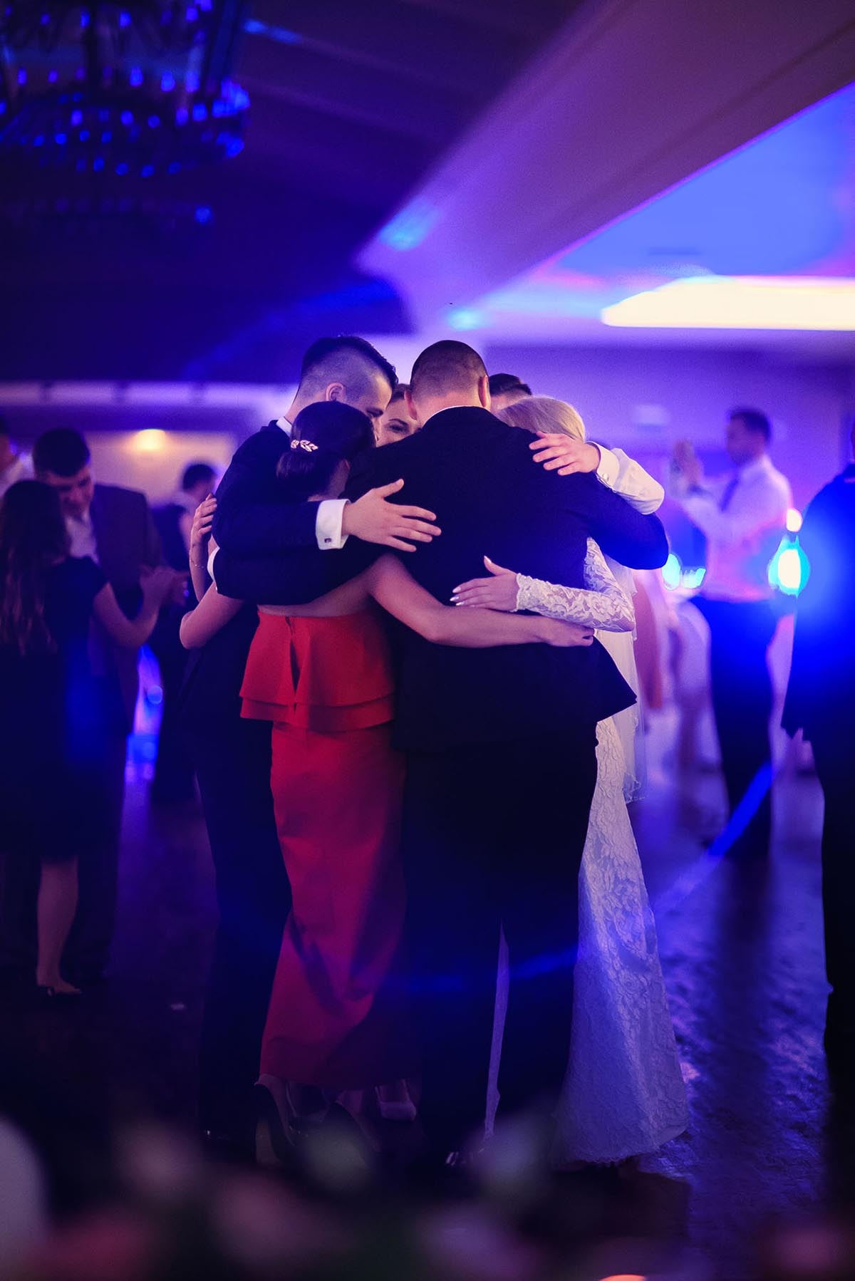 Uczestnicy wesela obejmują się podczas tańca w niebieskim oświetleniu sali. Widoczna panna młoda w białej sukni i goście w eleganckich strojach.