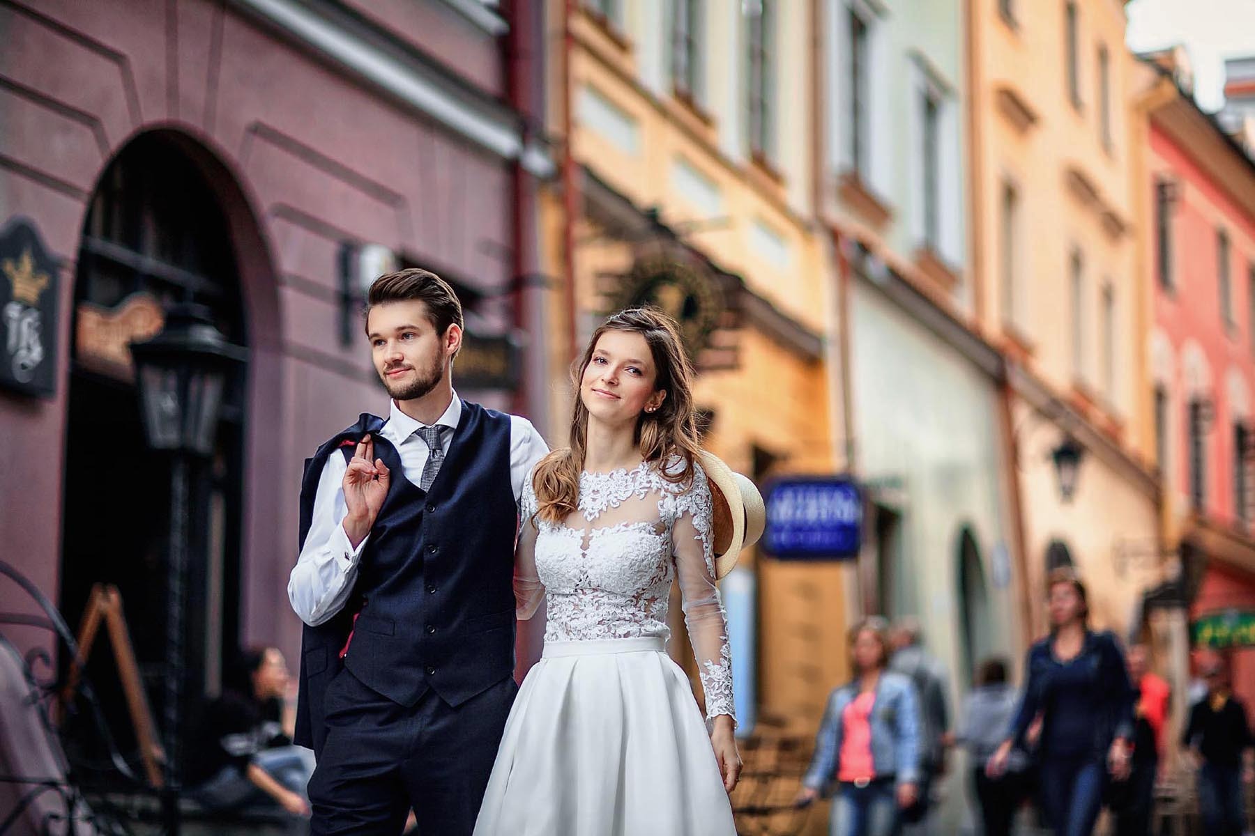 Para młoda spaceruje uliczką starówki. Pan młody w granatowym garniturze, panna młoda w białej sukni z koronką. W tle kolorowe kamienice i przechodnie.