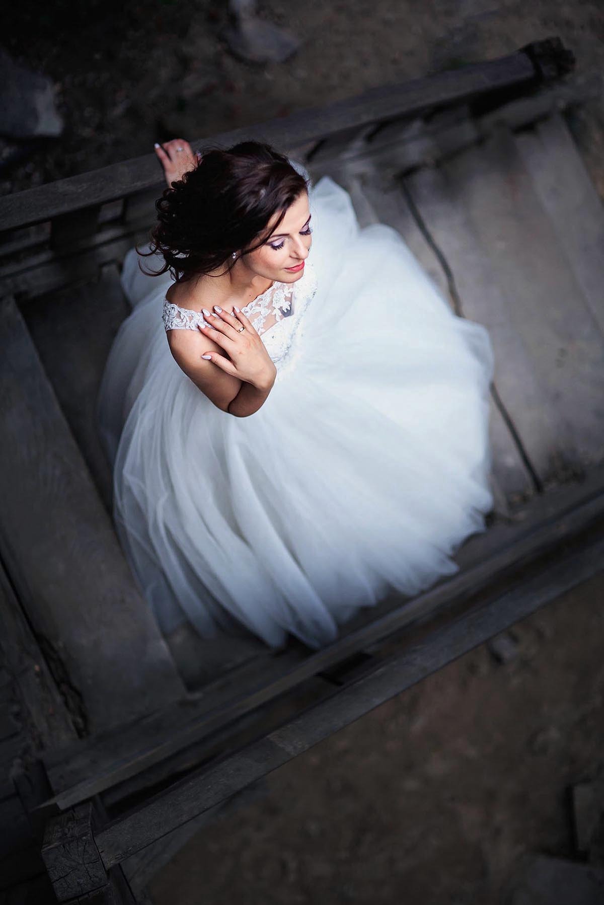 Panna młoda w białej sukni spogląda w dół, stojąc na drewnianych schodach. Ujęcie z góry podkreśla delikatność sukni i makijaż.