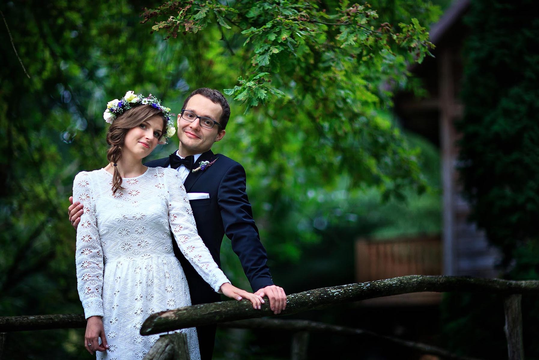 Para młoda w plenerze: panna młoda w koronkowej sukience i wianku, pan młody w eleganckim smokingu. Uśmiechnięci, opierają się o drewnianą barierkę na tle zieleni.