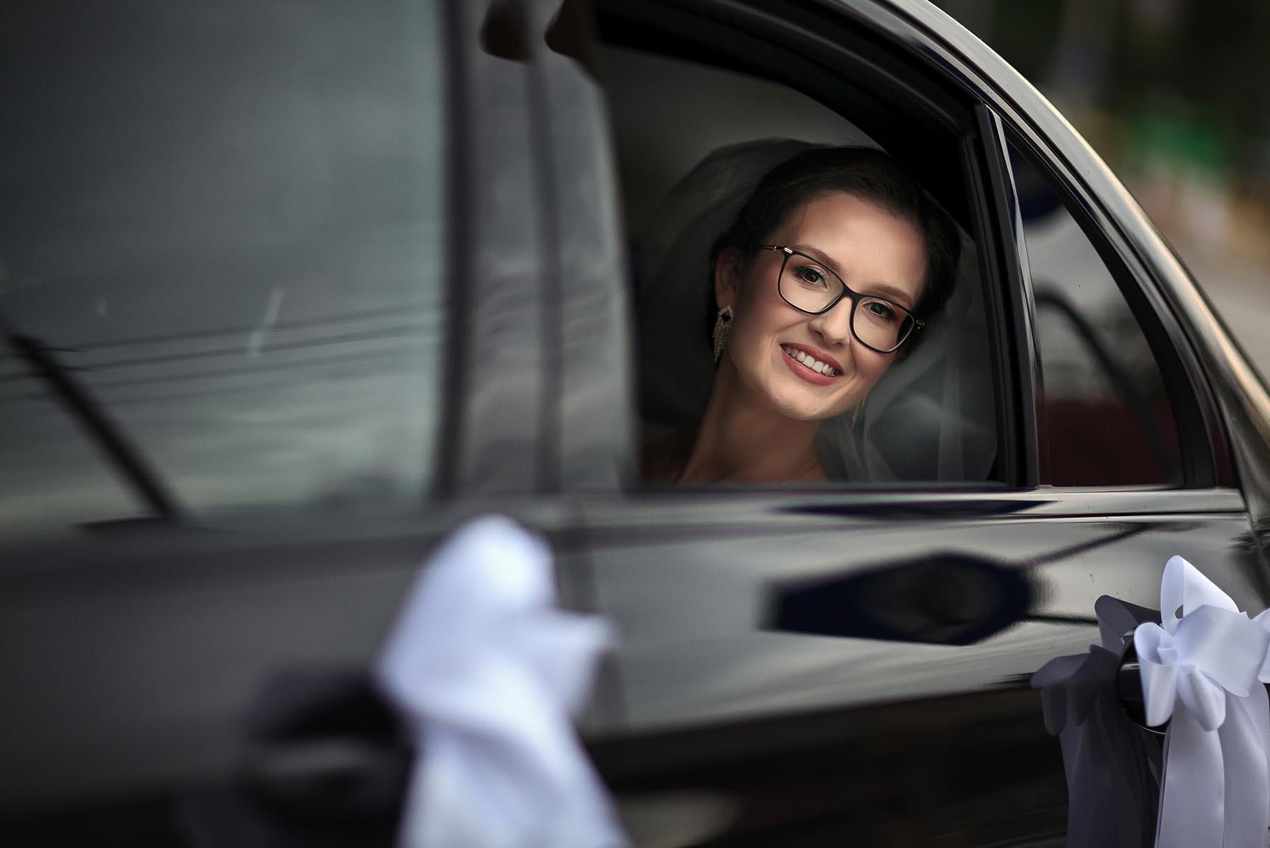 Uśmiechnięta panna młoda w okularach, spoglądająca z okna czarnego auta udekorowanego białymi kokardami, w dniu ślubu. Delikatny makijaż i welon.