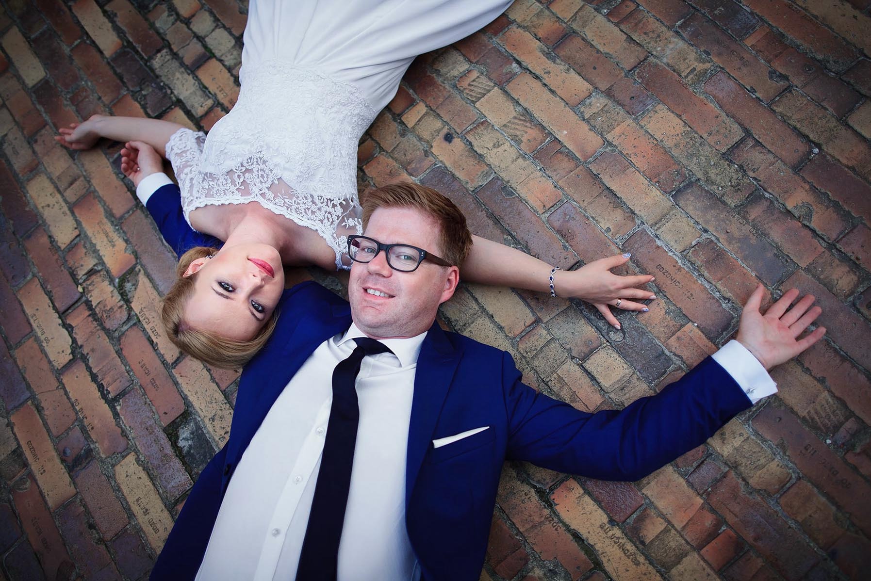 Para młoda leży na brukowanej powierzchni. Panna młoda w białej sukni z koronką, pan młody w niebieskim garniturze i okularach. Nietypowe ujęcie z góry, widoczne napisy na cegłach.