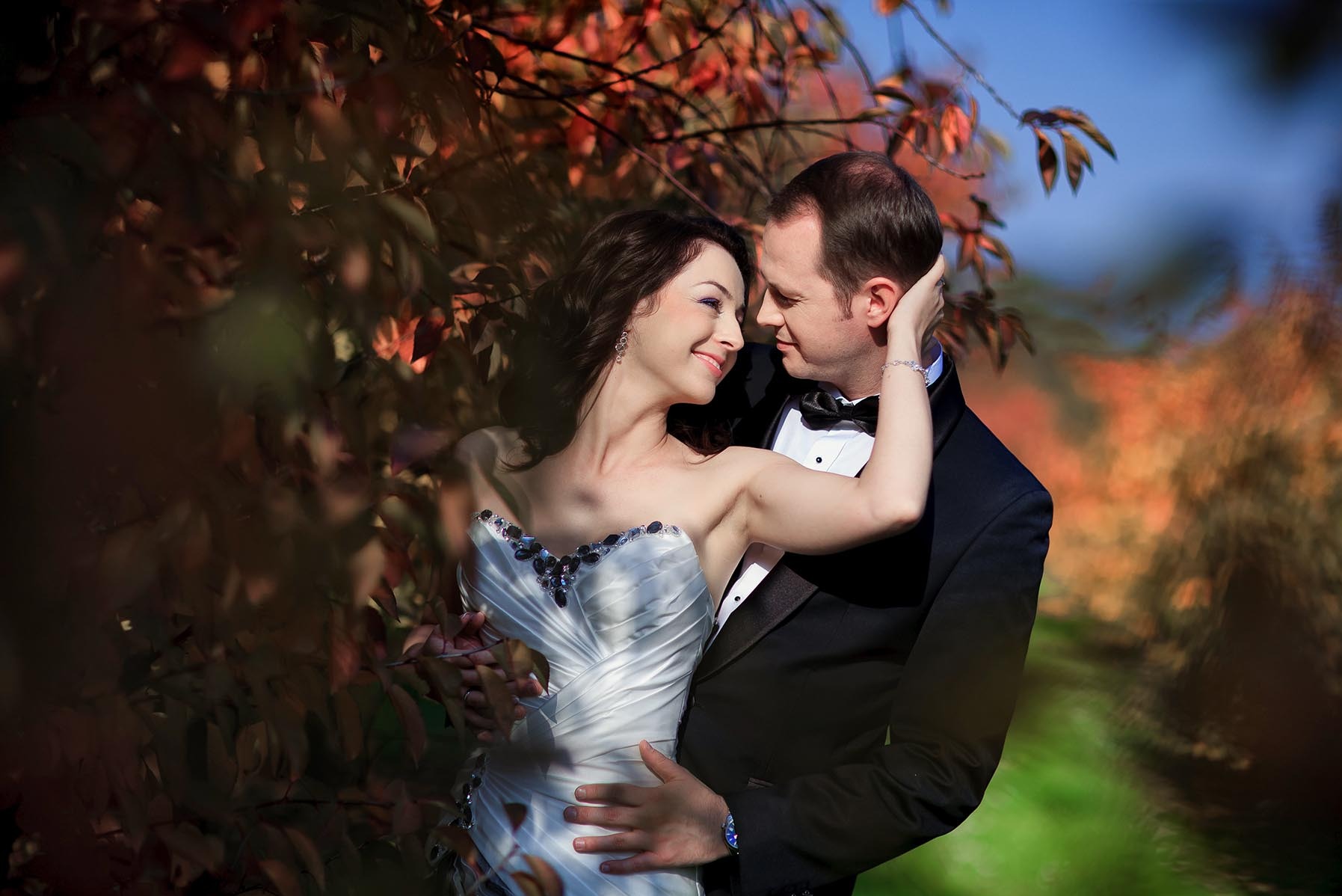 Uśmiechnięta para młoda w eleganckich strojach, otoczona jesiennymi liśćmi, w romantycznym ujęciu plenerowym z subtelnym, naturalnym światłem.