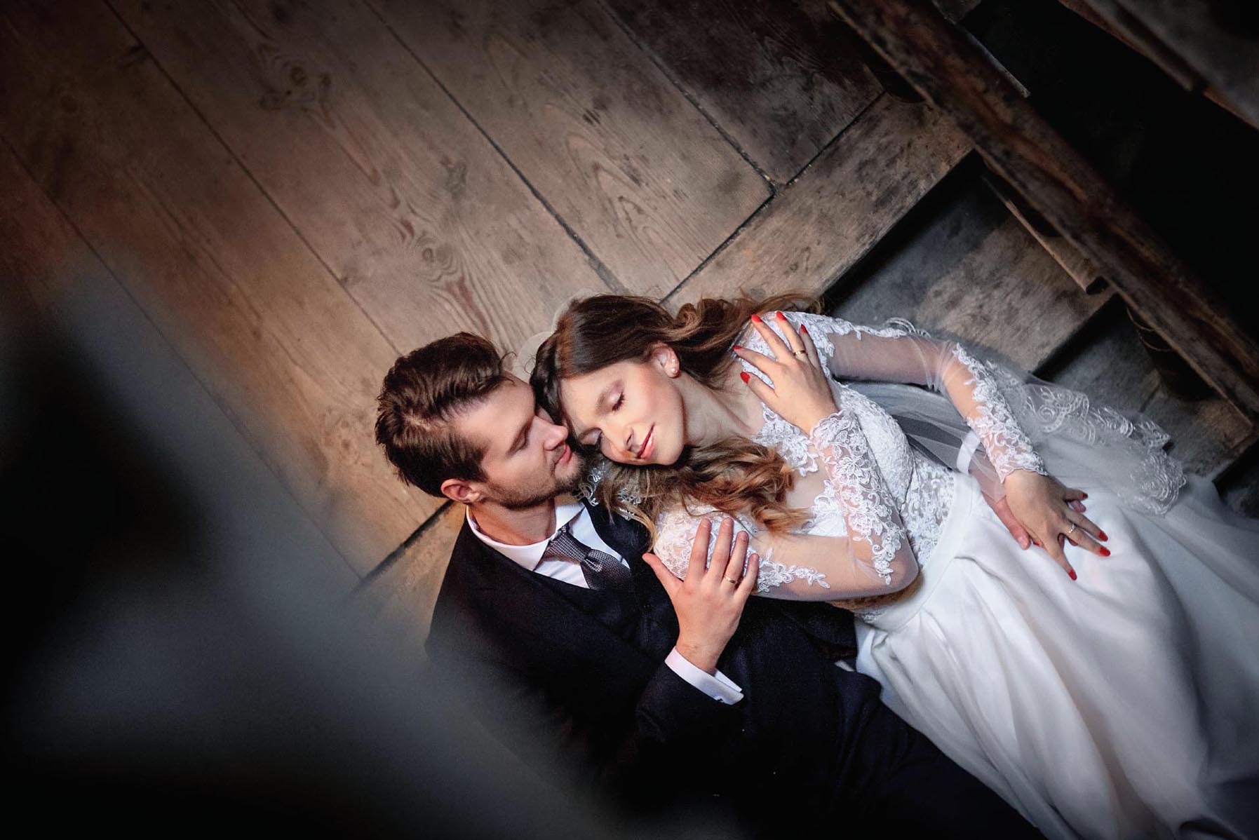 Romantyczne ujęcie ślubne: para młoda w objęciach na drewnianej podłodze. Ona w koronkowej sukni, on w eleganckim garniturze. Nietypowy kąt i ciemne tło dodają intymności.